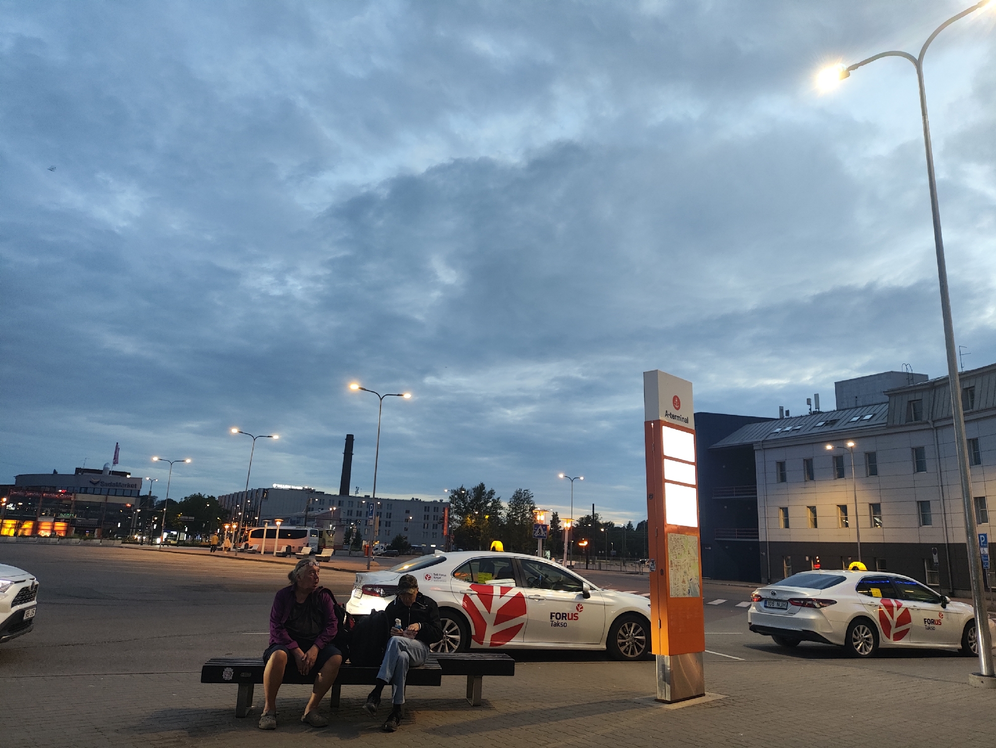 The image captures a street scene in Tallinn, Estonia, likely at a transportation hub given the presence of taxis and a terminal sign. In the foreground, two individuals are seated on a dark bench. One person, wearing a purple jacket and dark pants, appears to be looking off to the right. The other person, in a dark jacket and jeans, is holding a bottle and looking down at their phone.
Beside them are two white taxis, one with a "FORUS Takso" logo prominently displayed on its side. These taxis are positioned on a paved area, adjacent to a street where other vehicles, including a white car and a bus, are visible in the background. The environment suggests an urban setting with buildings lining the street, some featuring illuminated signage. Tall streetlights illuminate the scene, casting a glow against the backdrop of a cloudy, twilight sky. A prominent orange sign with "A-Terminal" text and illuminated panels stands near the taxis. The overall impression is that of a moderately busy public space during the evening hours.