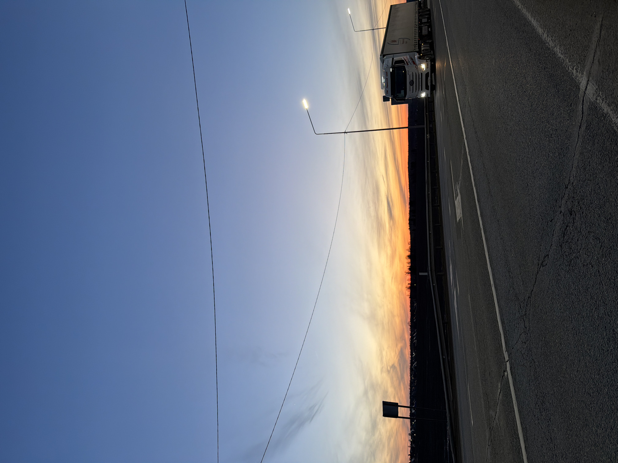 The image depicts a roadside scene at dusk or dawn, possibly in the vicinity of Kuregovo, Russia, given the presence of a truck with the text “ГЗК Пермскии край” (GZK Perm Region) visible on its side.
A large, white semi-truck is positioned on a multi-lane road. The road surface is asphalt and features lane markings. Streetlights illuminate the scene, one of which is positioned just before the truck.
The sky transitions from blue to orange/yellow, suggesting a sunset or sunrise. A strip of trees or vegetation is visible along the horizon. Power lines stretch across the sky in the left portion of the image.