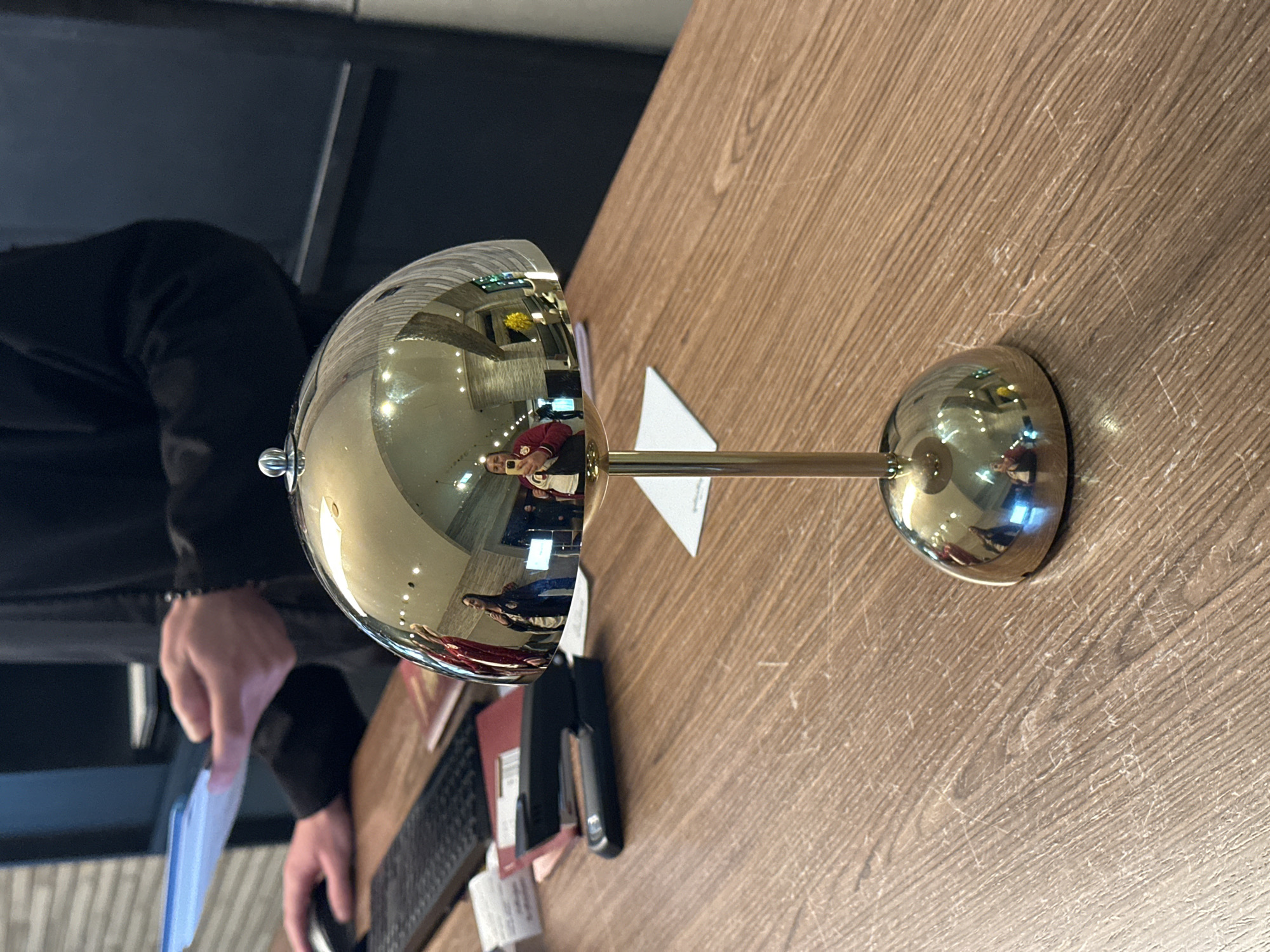 A golden desk bell sits on a wooden desk at what appears to be a hotel in Tbilisi, Georgia. A man in a black jacket is using a computer. In the reflection of the bell, one can see the lobby of a hotel, with a person wearing a red jacket.