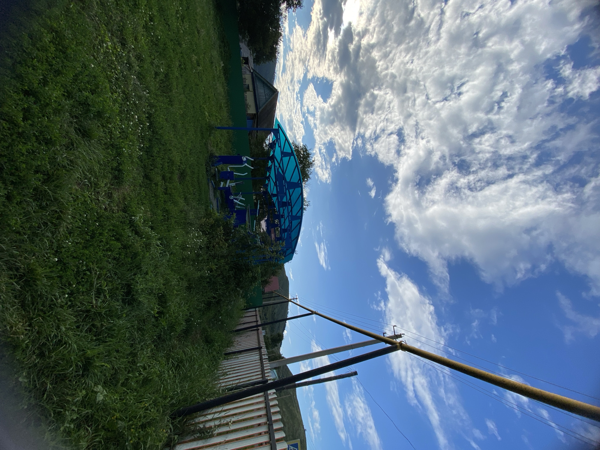 The image depicts an outdoor scene in Maloarslangulovo, Russia. In the foreground, a patch of green grass is visible, with some small white flowers scattered throughout. To the right, a white and rusted metal fence runs along the edge of the property. Beyond the fence, tall green grass and various plants grow. A large blue canopy structure with metal framing is situated in the mid-ground, covering several pieces of outdoor exercise equipment. A building with a brown roof and light-colored walls is visible behind the canopy and to the left, partially obscured by trees. In the background, rolling green hills rise against a bright blue sky filled with scattered white clouds. A prominent utility pole with wires stretches diagonally across the upper right portion of the frame.