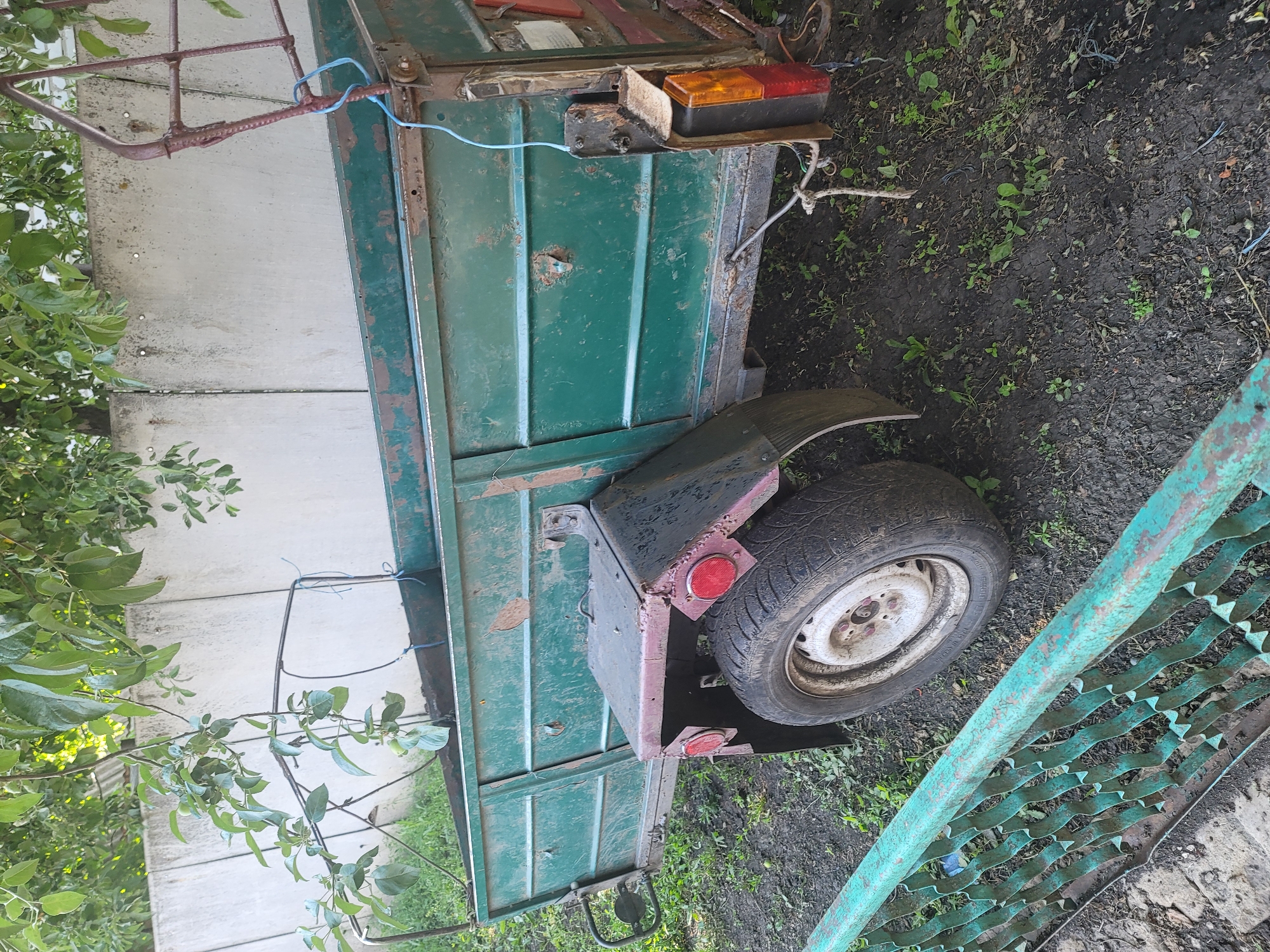 In the rural area of Vovkivka, Ukraine, an old, green, metal trailer is parked on uneven, muddy ground. The trailer, which appears to be weathered and shows signs of rust, has a single wheel visible on the right side. The wheel has a white rim and a worn tire. The trailer has a ribbed metal body and a dark metal fender over the wheel. A red and orange taillight assembly is attached to the rear right corner of the trailer. A rudimentary metal rack is positioned above the front of the trailer. Overhanging branches from a leafy tree partially obscure the top portion of the scene. In the background, a light-colored concrete wall runs horizontally, with some vegetation growing in front of it. A green, decorative metal fence runs along the bottom left of the image.