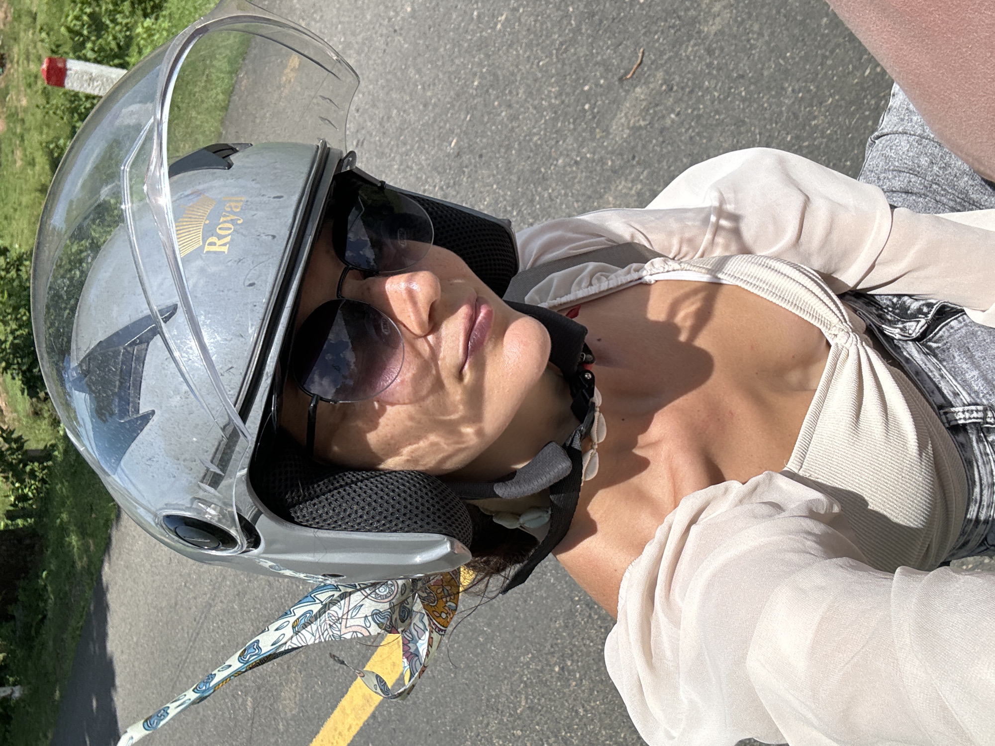 A woman wearing a grey helmet with a clear visor and sunglasses rides a scooter on a paved road. A patterned scarf is attached to the helmet. She is wearing a cream-colored top with puffed sleeves and grey shorts. The location is Ham Thuan Bac, Vietnam, a rural area with trees and vegetation visible in the background. The road has a yellow line marking the lane.