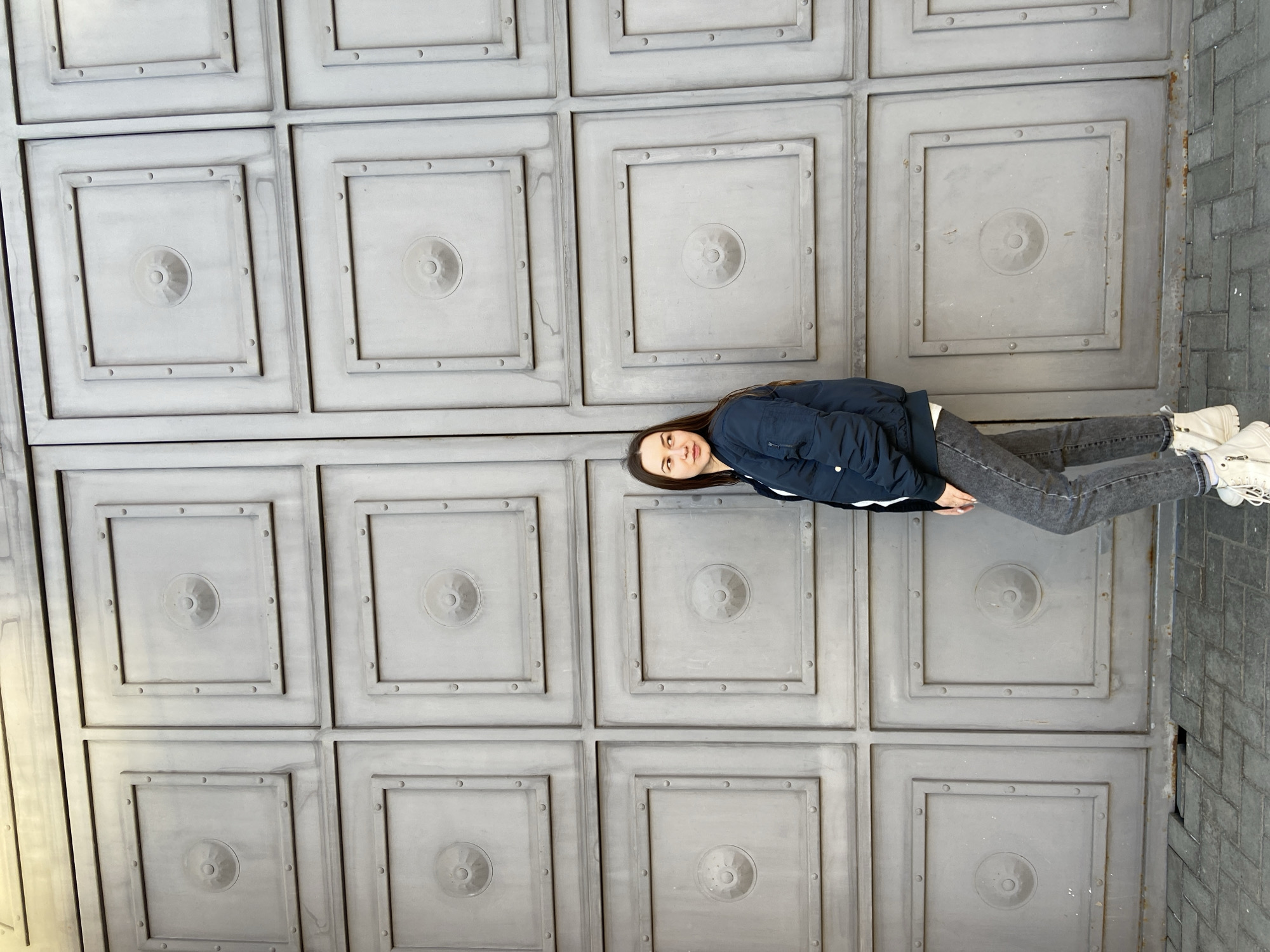 A young woman is depicted standing upright against a tall, grey, paneled wall or gate. The image, as provided, is rotated 90 degrees clockwise. The woman has long, dark hair and gazes directly at the camera with a calm, neutral expression. She is dressed in a dark blue bomber jacket, a white top, dark grey jeans, and light-colored boots.
The dominant background features a large, decorative structure crafted from numerous square panels arranged in a grid pattern. Each panel is distinguished by a raised square frame, accented with small, circular studs or rivets along its inner edge. A larger, concentric circular boss with a central recess adorns the middle of each panel. The structure's surface is a consistent grey, appearing to be made of metal or concrete, with subtle signs of weathering and discoloration.
To the woman's right and extending into the lower right of the frame, the ground is paved with grey rectangular bricks laid in a diagonal or herringbone pattern, indicating an outdoor setting such as a sidewalk or plaza. The lighting is soft and diffused, suggesting an overcast day or a well-lit shaded area, as evidenced by the absence of harsh shadows. There is no visible text within the image. The location context for this scene is Izhevsk, Russia.