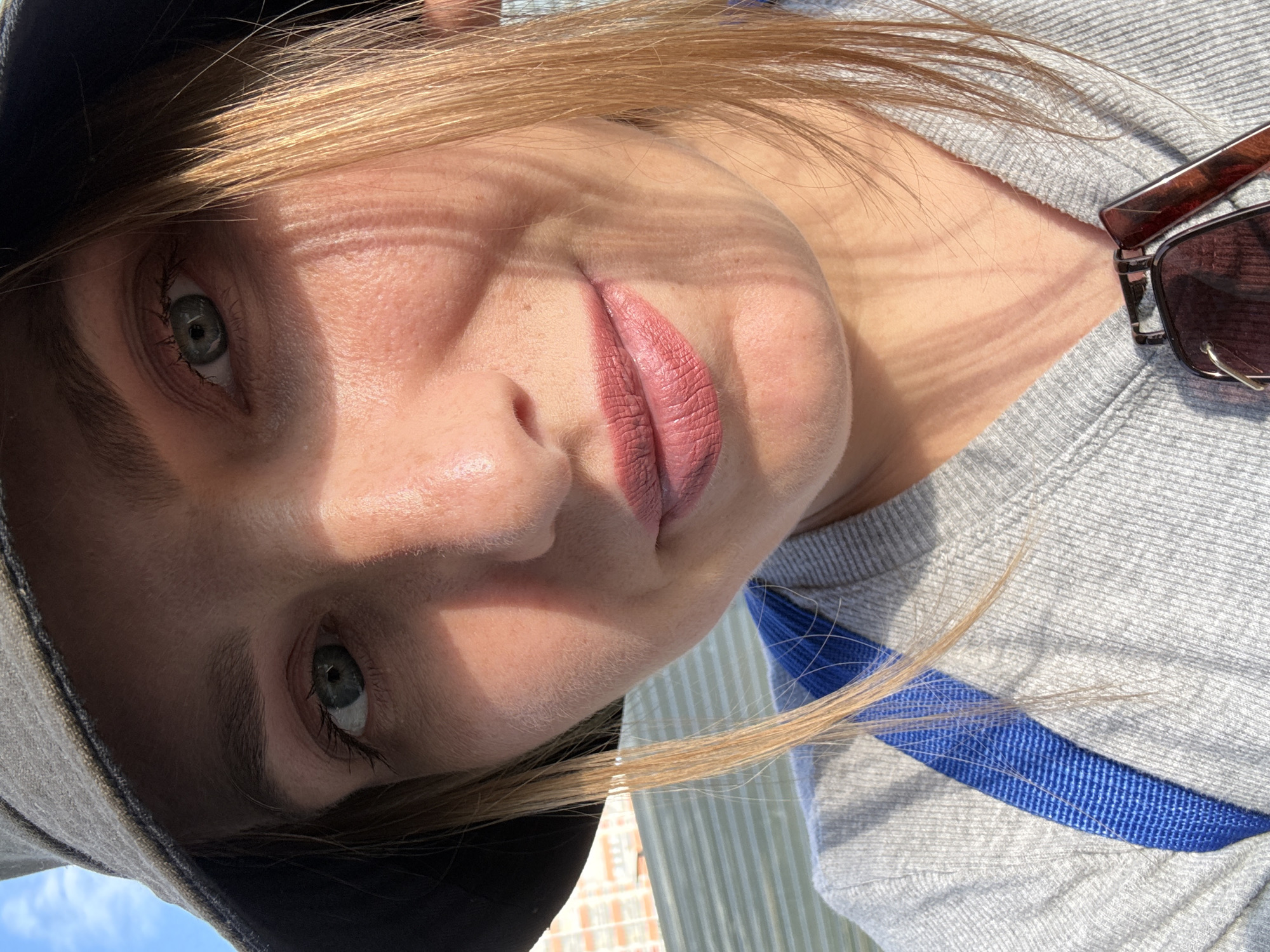 A woman wearing a grey bucket hat and a grey sweatshirt is taking a selfie outdoors. She has light skin, blonde hair, and blue-green eyes. Her lips are painted with a matte pink lipstick. There are shadows on her face from an unseen object, possibly foliage. A blue strap is visible across her chest, and sunglasses are clipped to her sweatshirt. In the background, a building with horizontal windows and a blue sky are visible. This appears to be a street scene in Kazan, Russia.