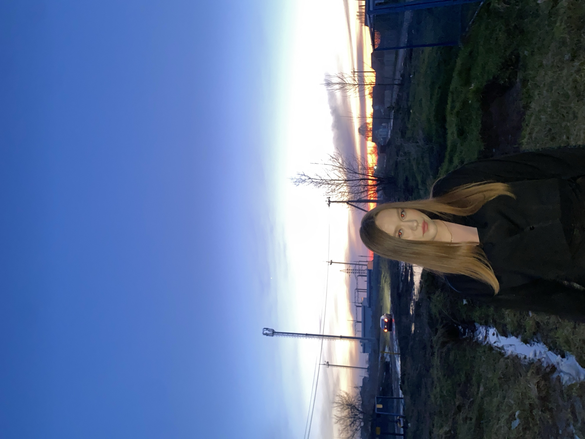 The image shows a young woman standing in an outdoor environment at dusk or dawn. The sky is a gradient of blue to orange, indicating either sunrise or sunset. In the background, there are utility poles and what appears to be an electrical substation. A vehicle's headlights are visible on the road. The foreground consists of grass and a puddle of water. The woman is wearing a dark-colored jacket.
The location is Chaikovsky, Russia.