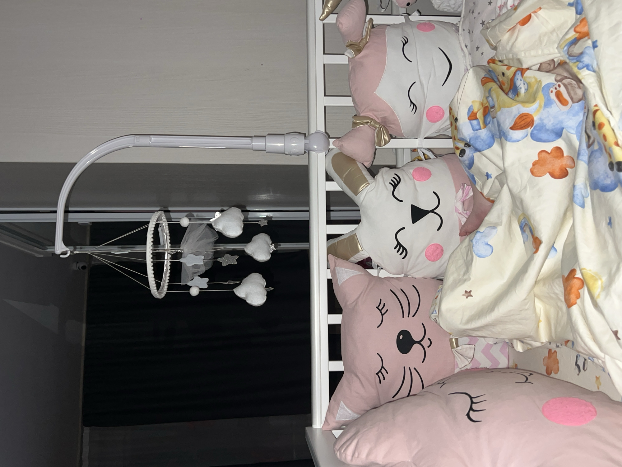 The image shows a baby crib with various soft toys inside. The crib is white with a mobile hanging over it. The mobile has small white cloud and star-shaped decorations. Inside the crib, there are several pink and white animal-shaped pillows, including a unicorn, a rabbit, and a cat. A patterned blanket with colorful designs, including giraffes, is draped over the pillows. The background shows a dark wall and ceiling. It is not possible to confirm the location in Chaikovsky, Russia, based on the visual information provided.