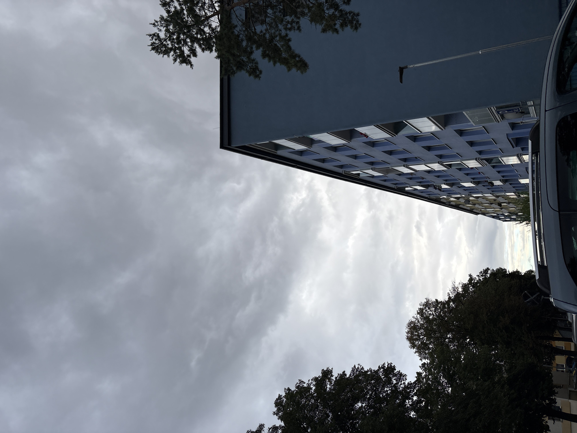 A low-angle, full shot captures a dark, cloudy sky over a blue apartment building and trees. The building, positioned on the right side of the frame, features numerous balconies with glass railings. A streetlamp stands tall on the building's right side. In the foreground, the roof of a dark grey vehicle is visible. On the left side of the image, a line of trees with dark foliage stretches across the frame, partially obscuring a row of buildings. The overall impression is one of a cloudy day in an urban setting.