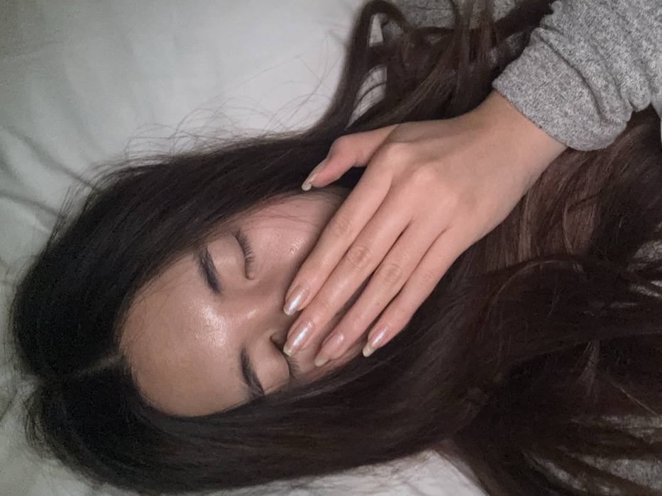 A close-up image depicts an individual, likely female, lying down with eyes closed. Her dark brown hair, parted centrally, spreads around her head. Her right hand, adorned with long, almond-shaped fingernails painted with a pearlescent polish showing visible regrowth, covers her nose and mouth. She wears a grey long-sleeved garment on her right arm. The background consists of a plain, light-colored, soft surface, consistent with bedding or a pillow, indicating an indoor setting.