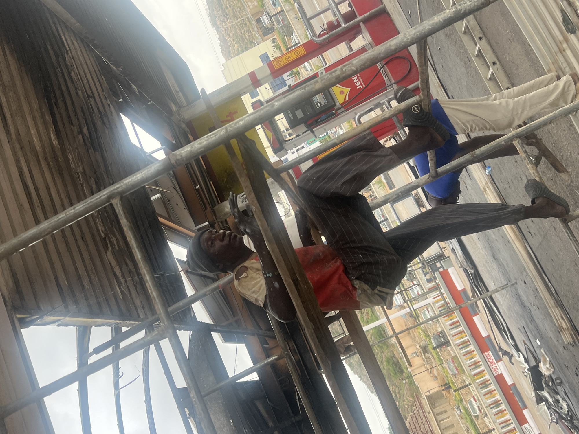 A man is working on scaffolding at a JENI petrol station in Gura Top, Nigeria. The man is wearing a white shirt, red covering, and striped pants. He is wearing sandals. The scaffolding is erected next to a damaged or dismantled structure with charred sections. In the background, there are buildings and a hillside. The petrol station has red and yellow branding. There is also a "no smoking" sign nearby.