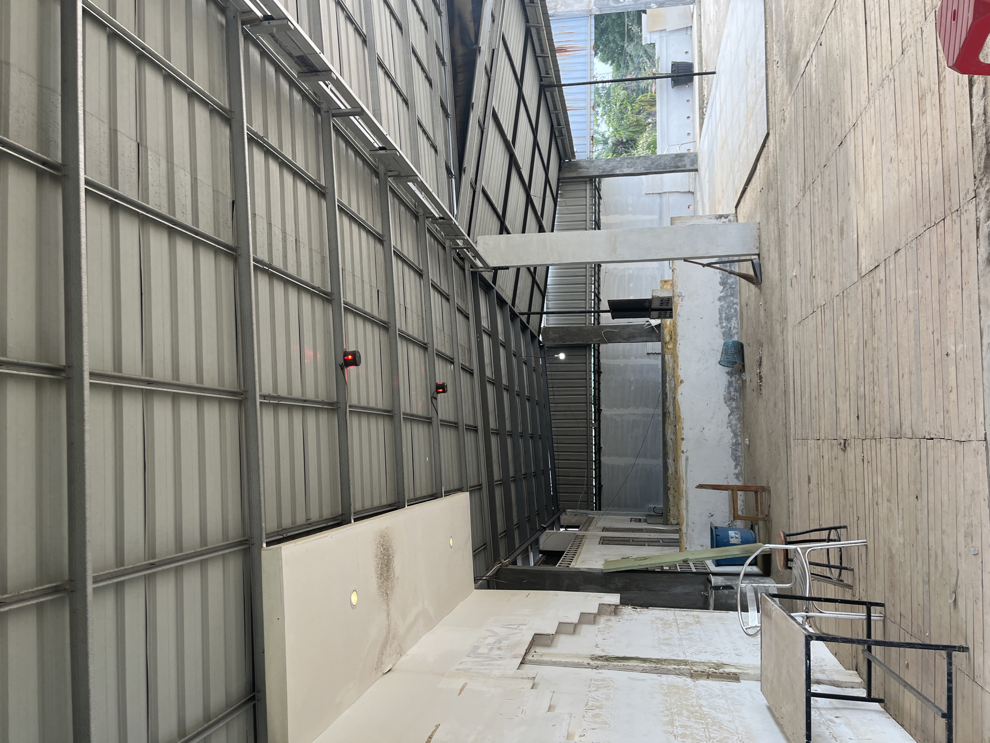 The image depicts the interior of an unfinished or under-construction building, likely a large open space such as a warehouse or commercial venue, located in Benuaraja, Indonesia. The dominant feature is the exposed metal roof structure with corrugated metal roofing sheets. The walls are partially finished, showing areas of bare concrete and some plastering.
In the foreground, there is a simple metal table with a wooden top, positioned on a wooden plank floor. Next to it are two chairs: a modern silver metal chair and a small wooden stool. Further into the space, more construction materials and debris are visible, including a blue plastic bin, a broom leaning against a pillar, and another wooden chair. The background reveals more of the building's structure, including concrete pillars supporting the roof, and what appears to be a more finished section of the wall. Beyond the open structure, trees and vegetation are visible, indicating an outdoor environment surrounding the building. The lighting suggests it is daytime, with natural light filtering into the space. There are no people present in the image.