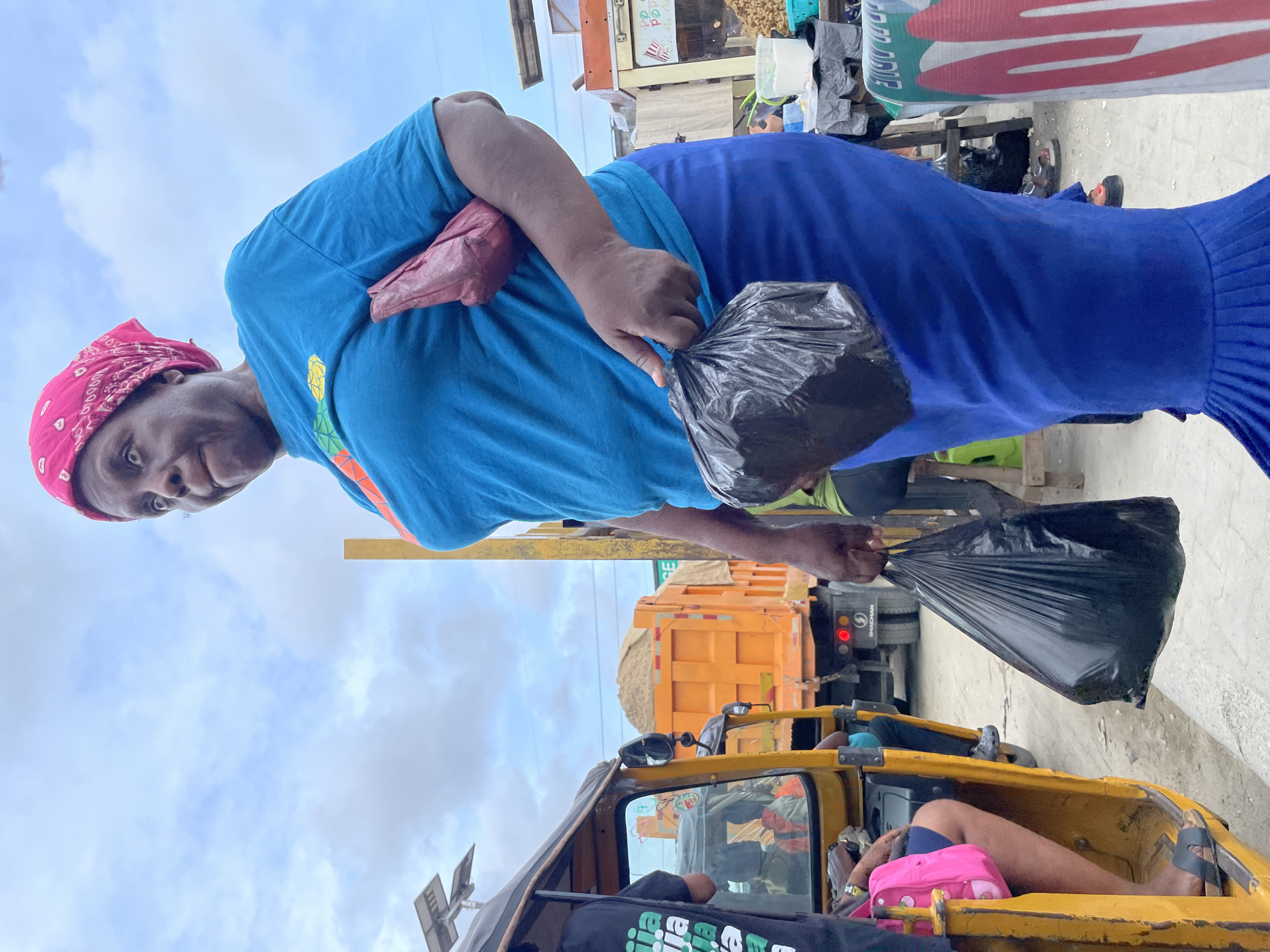 A medium shot captures a person standing outdoors, likely on a bustling street or market in Lagos, Nigeria, under an overcast sky. The person, with dark skin and a neutral to slightly smiling expression, is wearing a bright blue short-sleeved t-shirt with a colorful, pineapple-like graphic on the front, and a long royal blue pleated skirt or dress. A reddish-brown fanny pack or pouch is worn across their chest. They are holding two full black plastic bags, one in each hand, suggesting they are carrying purchases or goods. A vibrant red patterned headscarf completes their attire.
In the background, behind the person, an orange dump truck laden with sand is visible. To the right of the person, various market stalls or shopfronts can be seen, with a large white board or sign partially visible, displaying the text "GOOD TO DRIVE".
In the foreground, the top part of a yellow auto-rickshaw (locally known as a Keke NAPEP) is visible. Inside the rickshaw, a person's legs, clad in shorts and sandals, are seen, resting next to a pink bag. A black fabric or t-shirt with white and green text, possibly reading "BIG GEE", is also partially visible within the rickshaw. The overall scene depicts a typical daytime street environment, indicative of commercial activity and everyday life.