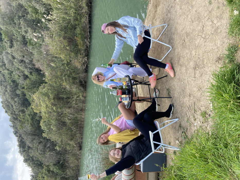 Four adult females are gathered outdoors near a body of water, possibly a river or canal, in the city of Unknown, Unknown. They are seated on portable folding chairs around a small, round, black folding table.
The woman on the far left wears a black long-sleeved shirt, a black puffer vest, black pants, and black sneakers. She smiles and holds a wine glass aloft in her raised right hand. To her right, a woman in a pink t-shirt, light-colored shorts, and a yellow long-sleeved garment draped over her shoulders, smiles and holds a stemmed glass in her raised left hand. On the center-right, a woman wearing a white long-sleeved top, white wide-leg pants, a light blue outer garment, and sunglasses is seated with her right hand resting near her head and her left hand on her lap; she is smiling. The woman on the far right wears a light blue denim jacket, dark pants, pink sneakers, and a pink baseball cap. She smiles and holds a green beer bottle aloft in her raised right hand.
On the table are a Coca-Cola bottle, two green beer bottles, two other clear bottles (one with a yellow label), a silver and white can, and a glass.
The ground beneath the group is dry, light brown soil with scattered green grass patches in the immediate foreground. Behind the subjects, a calm body of greenish-blue water flows, with its far bank densely covered in varied green foliage, including tall reeds, shrubs, and trees, extending up a gentle incline. The sky is overcast.
