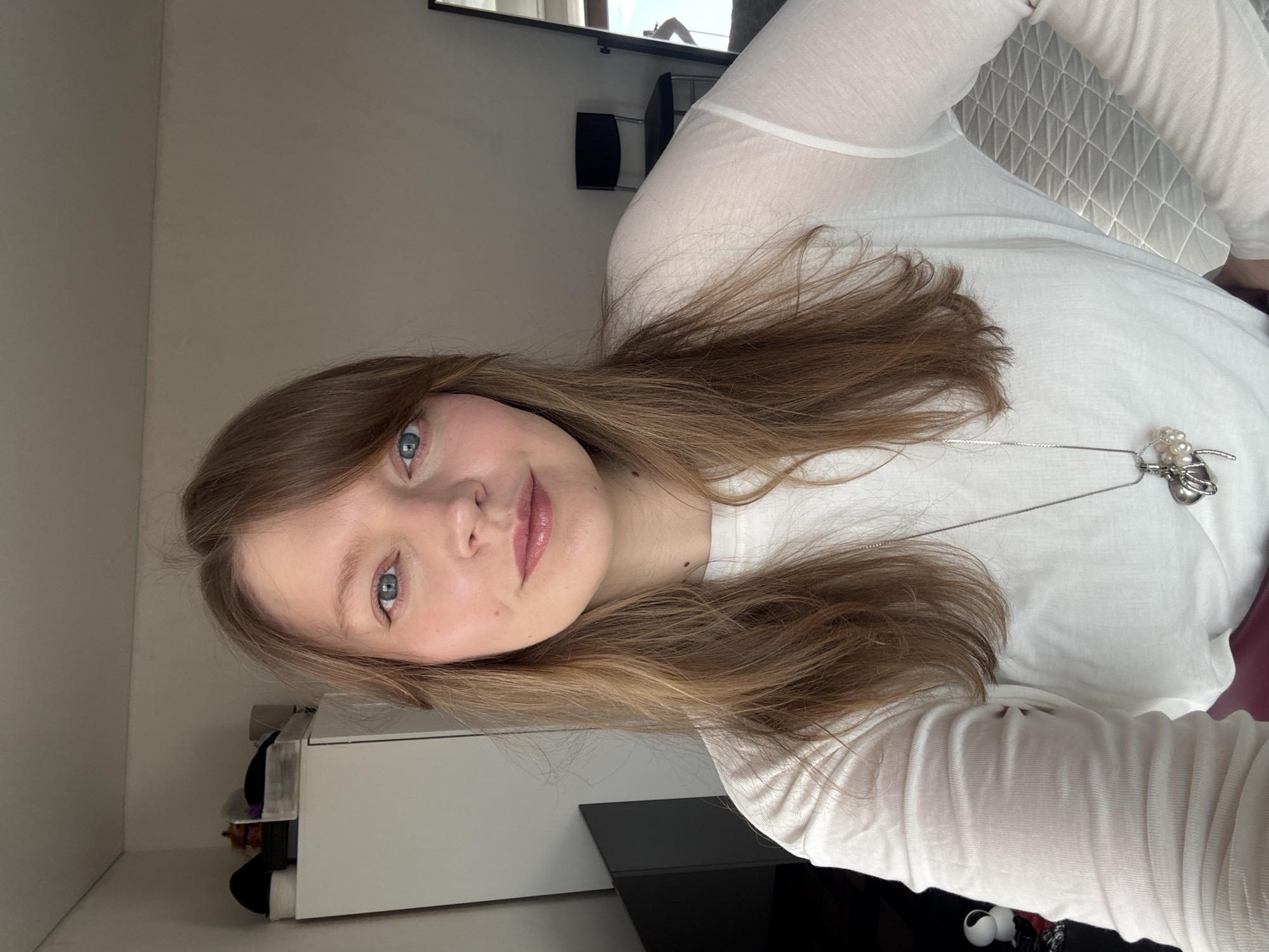 The image displays a head-and-torso selfie of a female individual captured indoors. The subject has long, light brown hair with bangs partially obscuring her forehead and blue eyes. She wears a sheer white long-sleeved top over an opaque white garment, and a silver-toned necklace with a pendant featuring a stylized metallic element and multiple white pearls. Her complexion is fair, and she has visible blush and lip tint. She looks directly at the camera with a neutral expression.
The background shows a domestic interior. To the viewer's left, a white wardrobe or cabinet is present, topped with several dark, indistinct objects. A dark, rectangular panel, possibly a television or monitor, is partially visible behind her left shoulder. To the viewer's right, a plain, light-colored wall extends, featuring a black minimalist chair with a metal frame. A tall, rectangular mirror is partially visible on the far right, reflecting a bright light source and a fragmented, obscured reflection of a person's lower torso. A grey, quilted or textured fabric is visible in the lower right portion of the frame, suggestive of bedding or upholstery.
The specific location within the city of Izhevsk, Russia, cannot be determined from the visual information provided in this image.