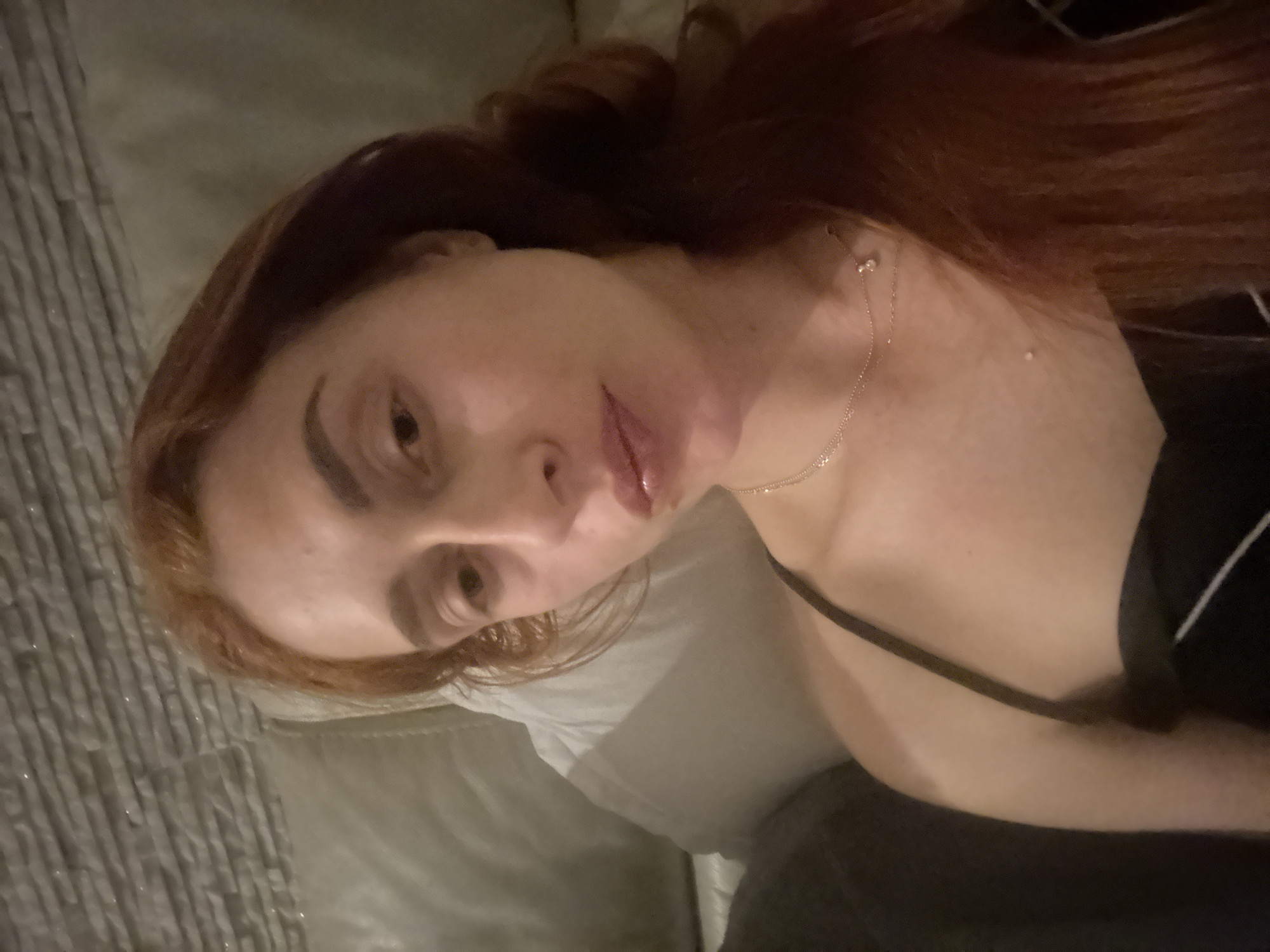 A woman with reddish-brown hair is reclining on a gray, textured cushion or bed. She is wearing a thin gold necklace and a black top with a thin strap. The background is blurred, preventing definitive identification of the location. No specific details indicate the city of Budva, Montenegro.