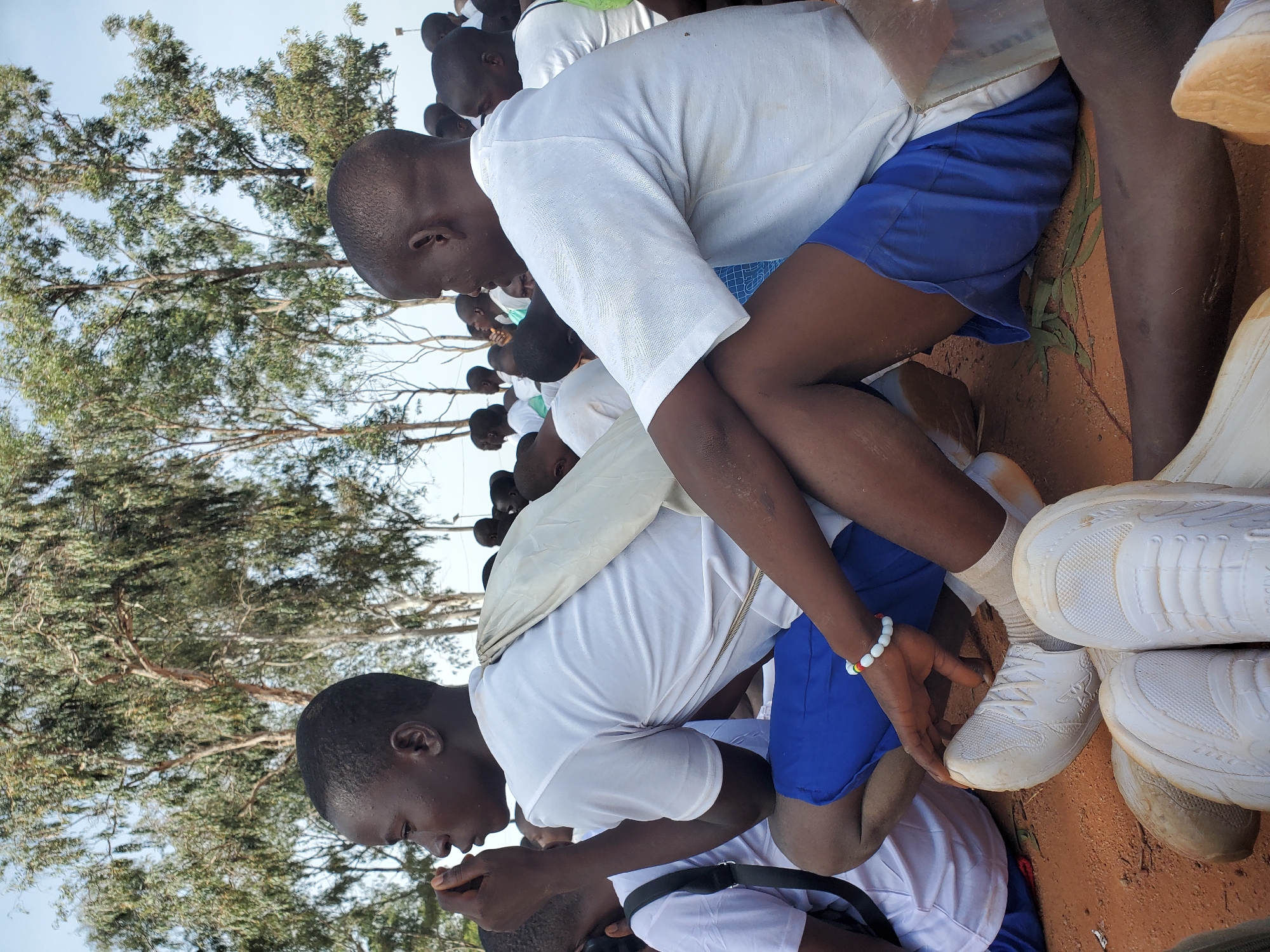 In Barakin Rafin Gora, Nigeria, a group of young people, likely students, are gathered outdoors, sitting on the ground. The foreground is dominated by the legs and feet of several individuals. Two young men in the center are the most prominent subjects. One is wearing a white t-shirt and blue shorts, with a white bag slung over his shoulder. He is looking to his left, with his right hand near his mouth. He has a bracelet made of colorful beads on his left wrist. The other young man, to his right and slightly behind him, also wears a white t-shirt and blue shorts. His head is turned to his left, and his right arm rests on his bent leg. Both are wearing white sneakers. In the background, more individuals in similar attire are visible, sitting and standing. The environment appears to be a dry, sandy area with sparse vegetation and tall trees with green foliage overhead. The lighting suggests it is daytime.