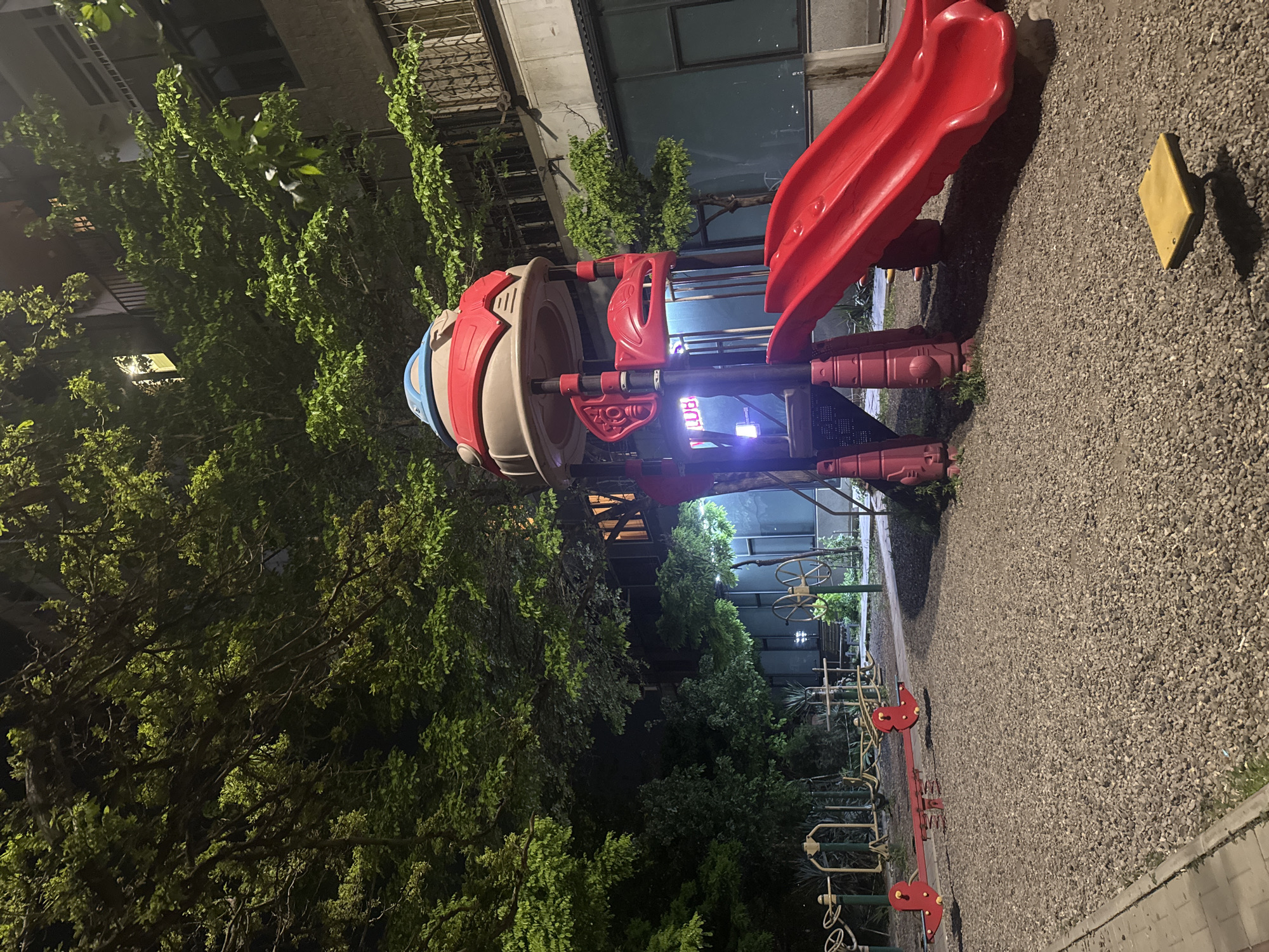 The image captures a children's playground in Yerevan, Armenia, at night. The playground equipment includes a red slide attached to a spaceship-themed structure with a beige body and red and blue accents. Other playground elements, such as a seesaw, are visible in the background. The ground is covered in gravel. The playground is surrounded by lush greenery, including trees and shrubs, contributing to the nighttime ambiance. A building is partially visible behind the playground, suggesting an urban setting.