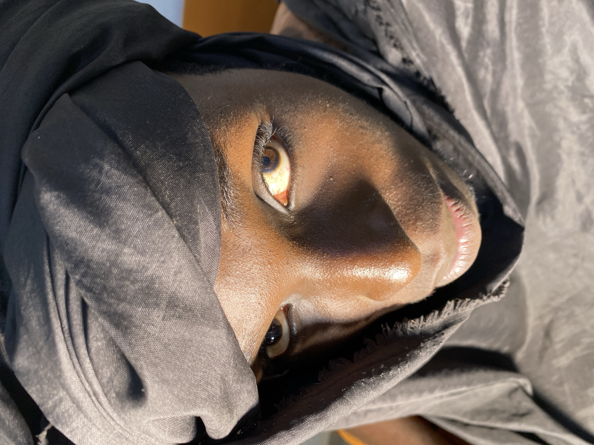 A close-up portrait shows a young person wearing a black turban and clothing. Their face is turned upwards, and the sunlight highlights their features. The person has dark skin and brown eyes. The fabric of the turban is draped around their head, partially obscuring their face. The background is out of focus, but appears to be an indoor setting with a warm-toned surface, possibly a wall or furniture.
