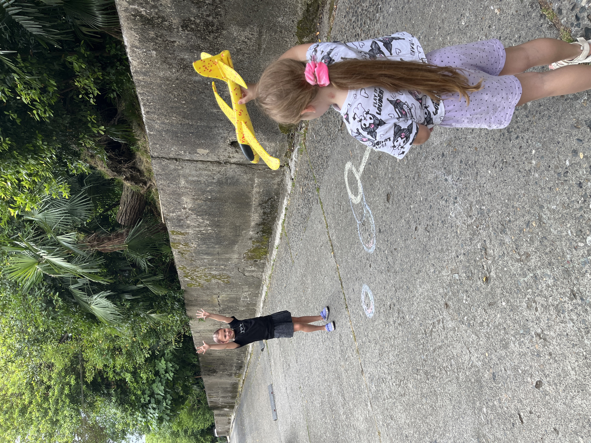 Two young girls are playing with a yellow foam airplane on a paved road in Sochi, Russia. The girl on the left, wearing a black t-shirt and grey skirt, stands with her arms raised and palms outward. The girl on the right, facing away from the camera, holds the airplane in her right hand and wears a white t-shirt with black panda illustrations and purple shorts. The background features a low concrete wall and lush green foliage, indicative of the subtropical climate of Sochi. Chalk drawings of circles are visible on the asphalt in front of the girls.