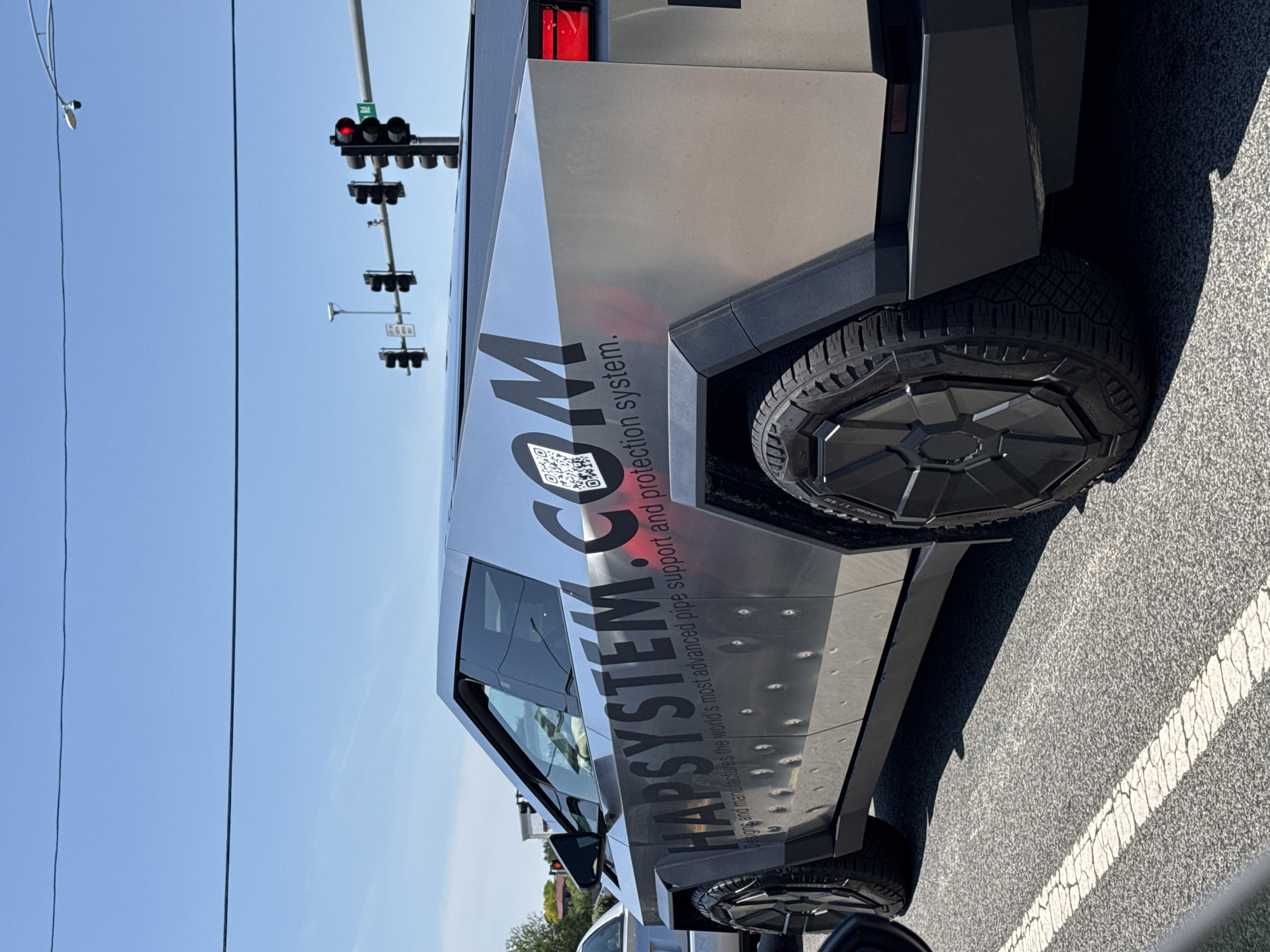 A Tesla Cybertruck is parked on a street in Glendale Heights, Illinois, United States of America. The Cybertruck has "HAPSYSTEM.COM" printed on its side along with a QR code. A traffic light is visible in the background. The sky is clear.