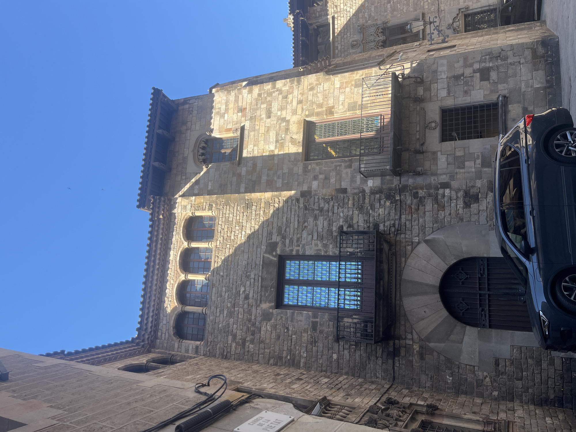 The image depicts a view of a stone building in Barcelona, Spain, likely in the Gothic Quarter. The architecture features stone walls, arched windows, and balconies with iron railings. A car is parked on the side of the street next to the building. There's a street sign visible with the name "Carrer de la Pietat". The sky is clear and blue. The building has areas that are brightly lit by sunlight and others that are in shadow, possibly indicating the time of day.
