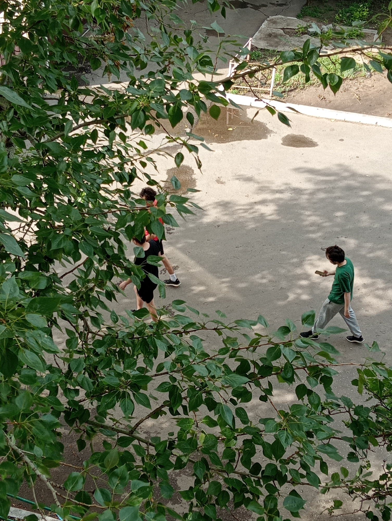 The image shows a courtyard with two individuals walking. One individual, wearing a green t-shirt and gray pants, is walking away from the camera while looking at a mobile phone. The other individual, wearing a black t-shirt and shorts, is walking towards the camera, with another person in a red t-shirt and black shorts walking in the same direction but slightly ahead. They appear to be on a paved surface with some patches of wetness and shadows from overhead foliage. The scene suggests a casual, outdoor setting, possibly a residential area given the presence of a low concrete structure and greenery. The image context is a courtyard in Kostanay, Kazakhstan.
