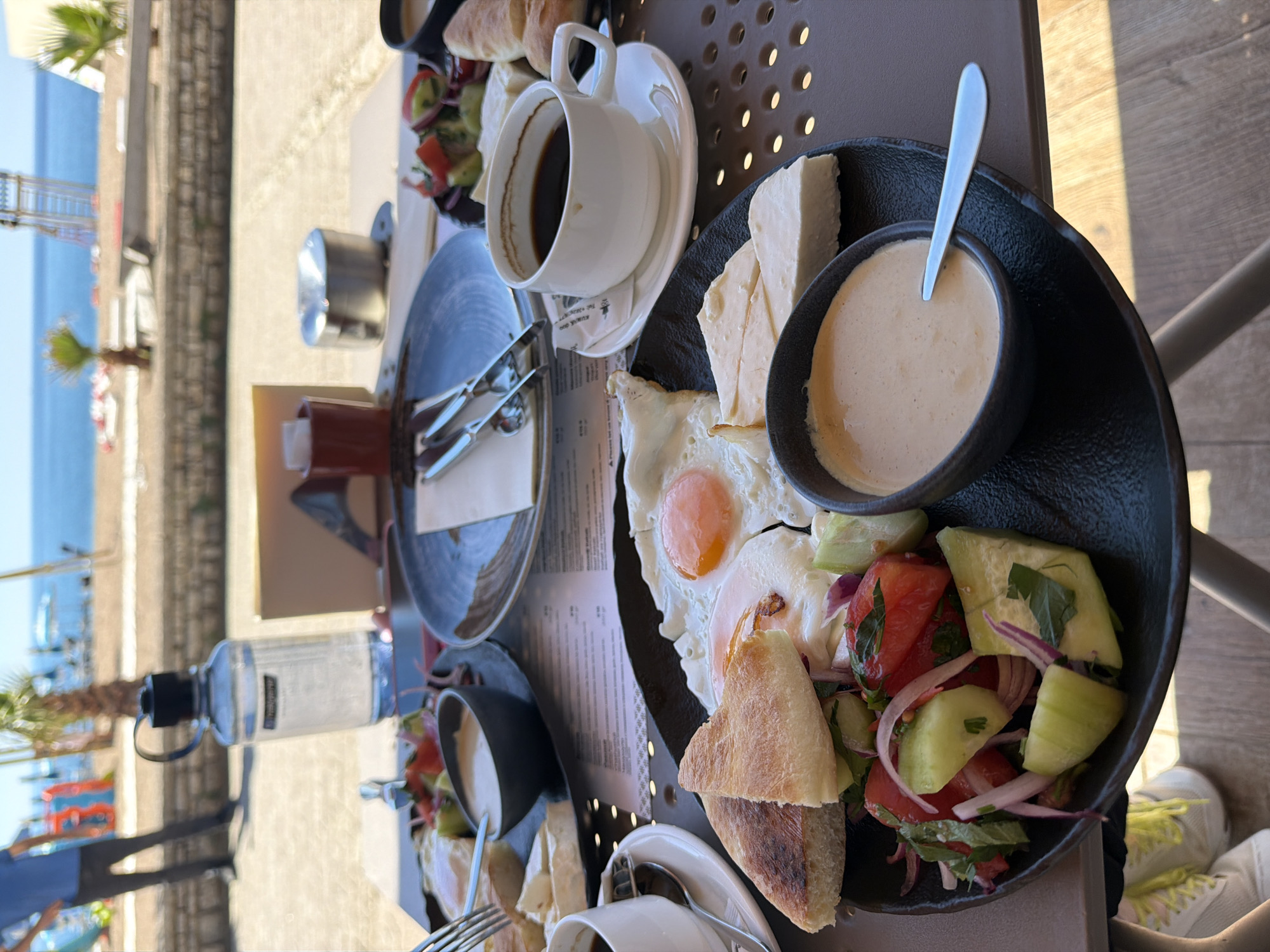 A breakfast scene is captured in Boreti, Montenegro, revealing a table laden with food against a backdrop of the sea and coastline. Two breakfast platters are prominent, each containing fried eggs, salad with cucumbers and tomatoes, white cheese, and a bowl of sauce. Bread accompanies the meal. Two cups of coffee are present. A water bottle rests on the table near a menu. The overall setting suggests an outdoor breakfast experience with a view of the coastal city.