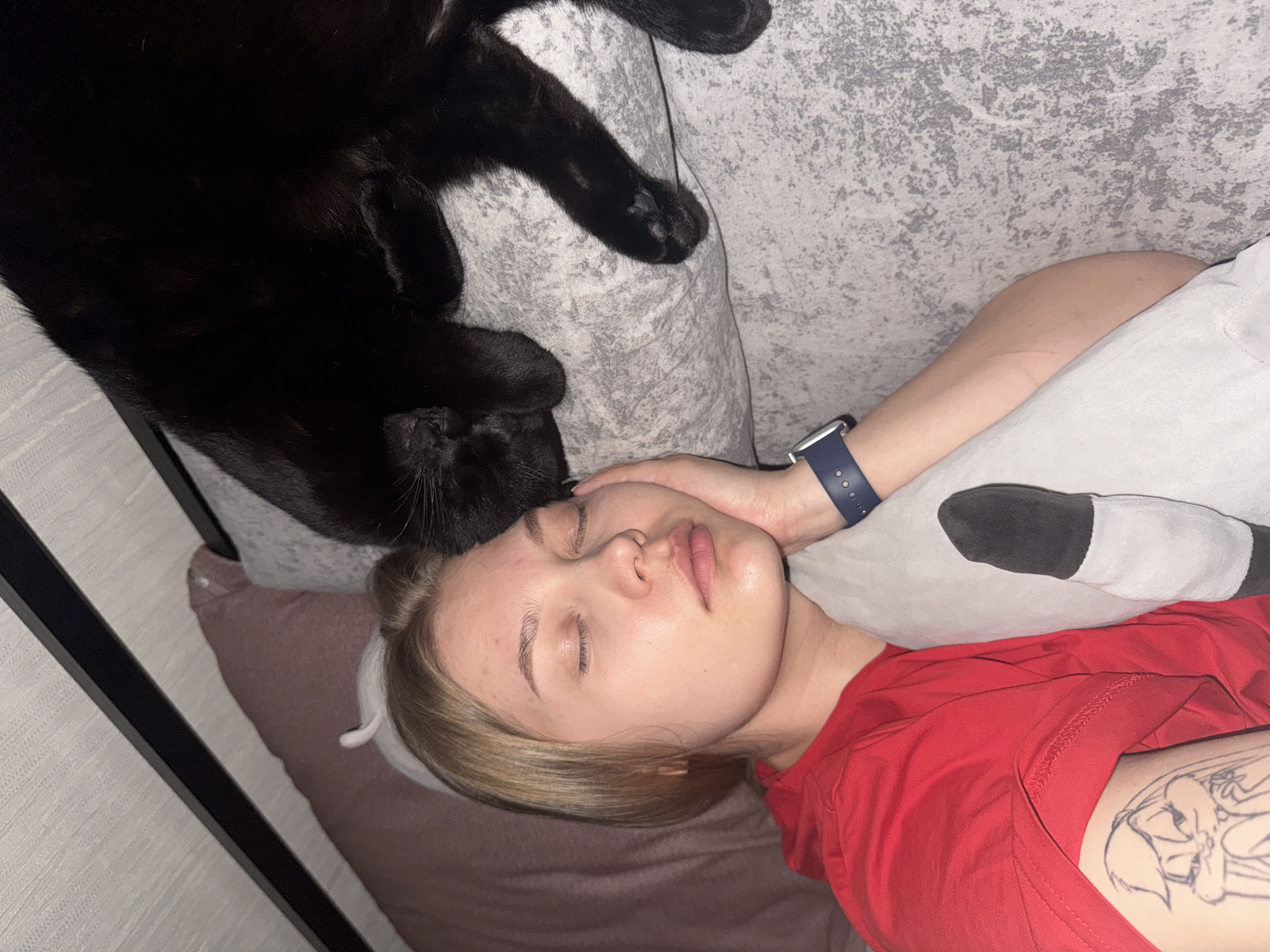 A photograph, likely taken in a residential setting, depicts a woman lying down with a black cat. The woman is fair-skinned, blonde, and wearing a red t-shirt. A tattoo is visible on her upper arm. She has a blue smartwatch on her wrist. She is lying on a grey patterned couch and grey/white pillow. The black cat is resting on the pillow near the woman's head. A black doorframe is visible on the left side of the photo. The location is not ascertainable from this image.