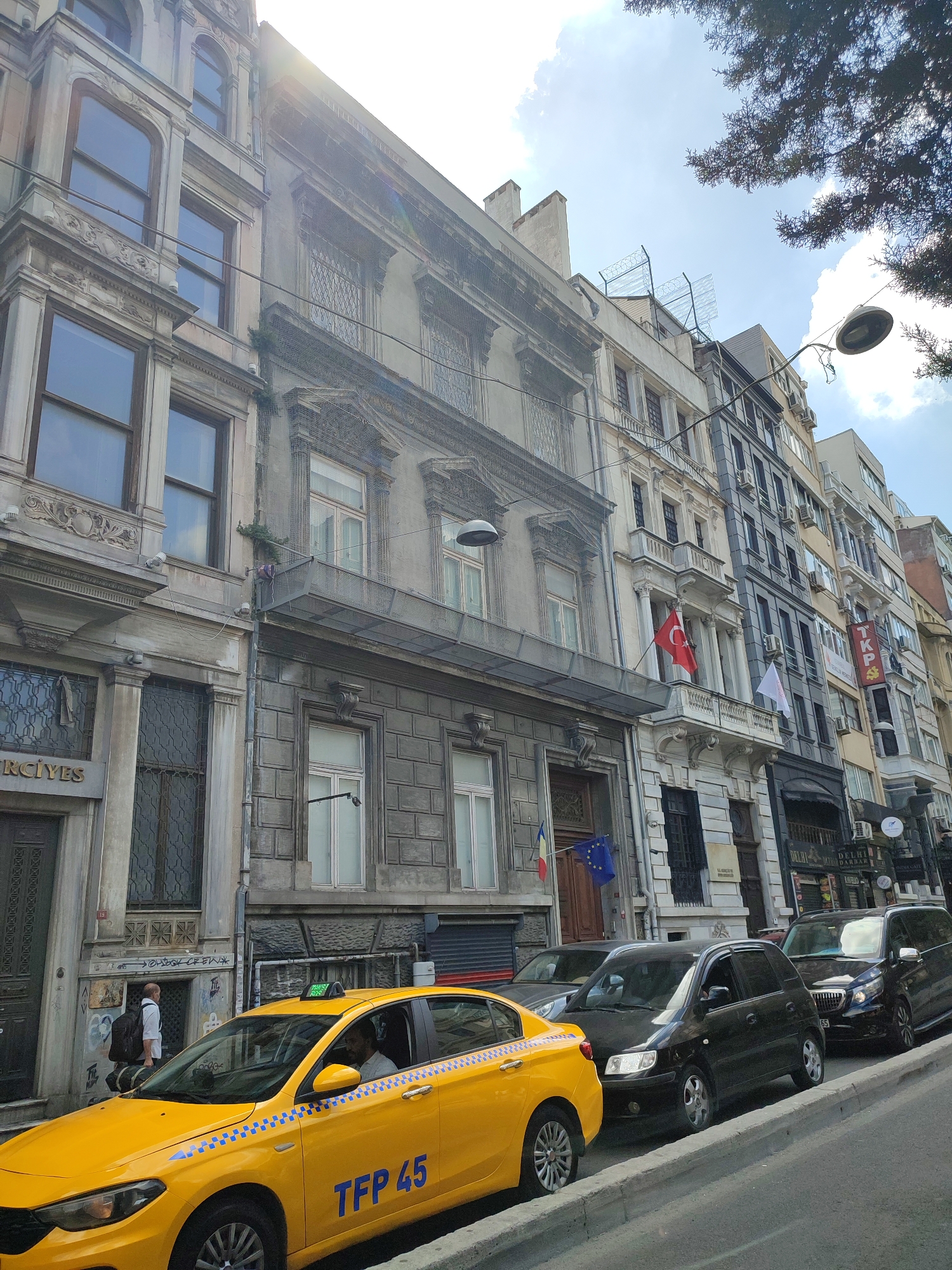In Beyoğlu, Turkey, a street scene unfolds with several vehicles lined up on the right side of the image, predominantly dark in color, including a black minivan and a black sedan. A vibrant yellow taxi with the license plate "TFP 45" is the most prominent vehicle, positioned in the foreground on the left. A person wearing a white shirt and carrying a dark backpack is walking away from the camera towards the left side of the image, in front of a building. The buildings lining the street exhibit varied architectural styles, with some featuring ornate facades and balconies. Flags are visible on some of the buildings, including a Turkish flag and an EU flag. The sky is partly cloudy, and sunlight casts shadows on the buildings and street. The overall impression is a bustling urban environment with a mix of historical and modern elements.