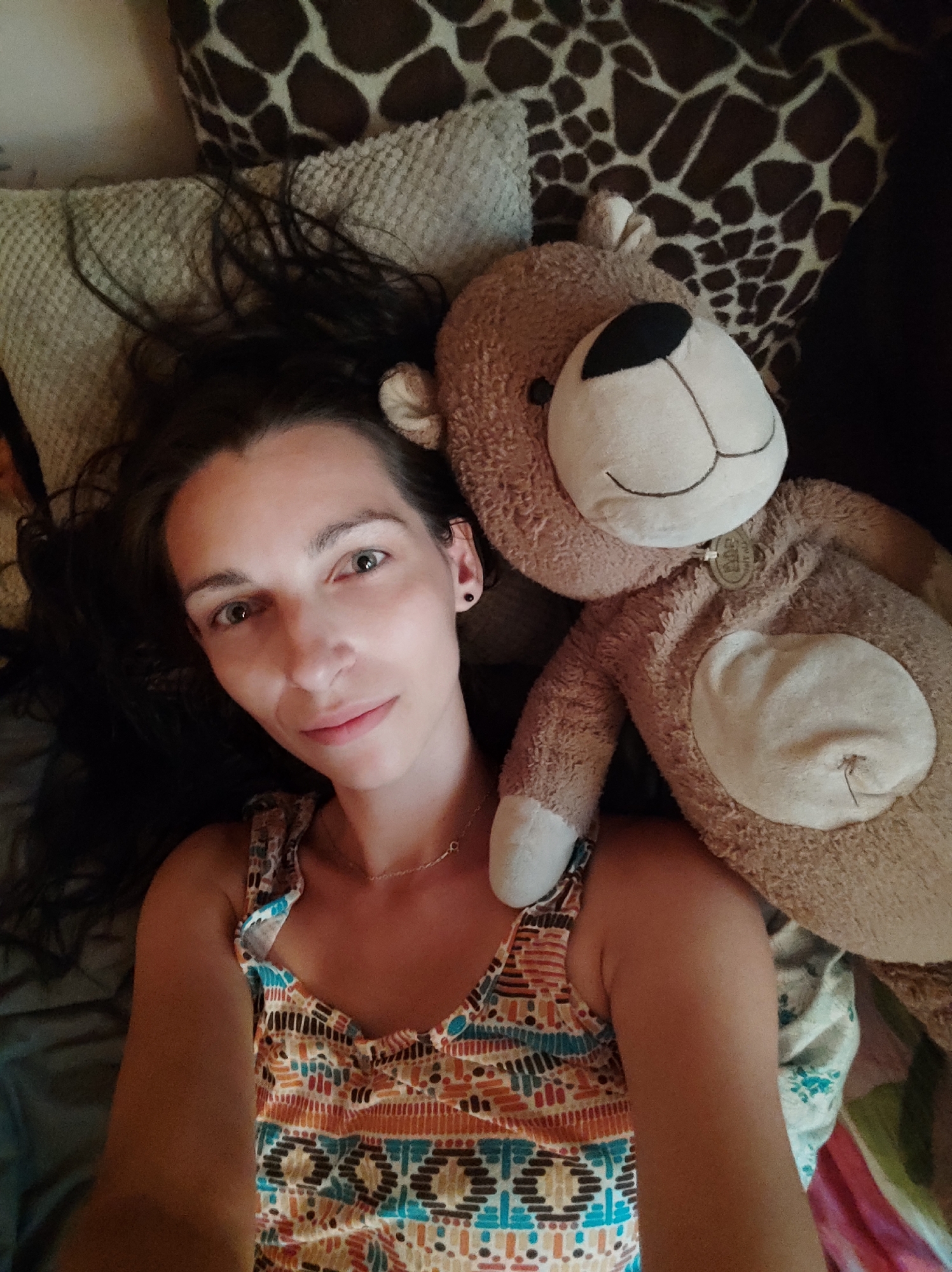 The image shows a top-down selfie of a woman with dark hair lying down next to a teddy bear. The woman is wearing a colorful tank top with a geometric pattern and a thin necklace. Her head is tilted slightly to her left, and she is looking directly at the camera with a faint smile. Her left arm is bent and resting on her chest, while her right arm is obscured by the teddy bear. The teddy bear is large and light brown, with black embroidered eyes and a stitched smile. It has a circular patch on its chest. Behind the woman and the teddy bear, there are pillows with patterned covers, one with a giraffe print and another with a textured weave. The setting appears to be indoors, possibly a bedroom, given the presence of pillows and bedding. There is no identifiable information in the image to suggest it was taken in Kryvyi Rih, Ukraine.