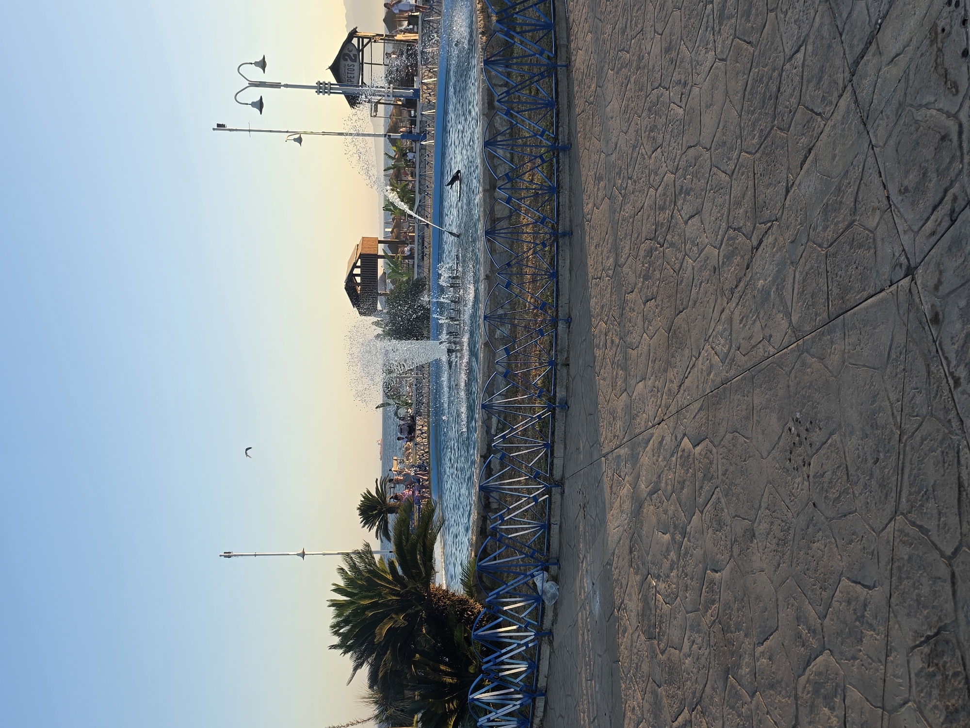 The image depicts a public space with a fountain in the foreground, set against a backdrop of a clear, light blue sky transitioning to a pale yellow horizon. The fountain features multiple water jets and a dark, elongated object, possibly a decorative statue or a seal, partially submerged in the water. Surrounding the fountain is a blue metal fence with a diamond pattern. Beyond the fence, palm trees are visible, along with a paved area with large, irregularly shaped stones. In the background, there are several small structures with thatched roofs, possibly kiosks or beach shelters, and people are seen walking or sitting. The sea is visible in the distance, with a few ships on the horizon. A lamppost stands to the left of the fountain, and another lamppost with multiple lamps is located to the right, near one of the thatched structures. A bird is flying in the sky. The overall scene suggests a seaside resort or promenade area.