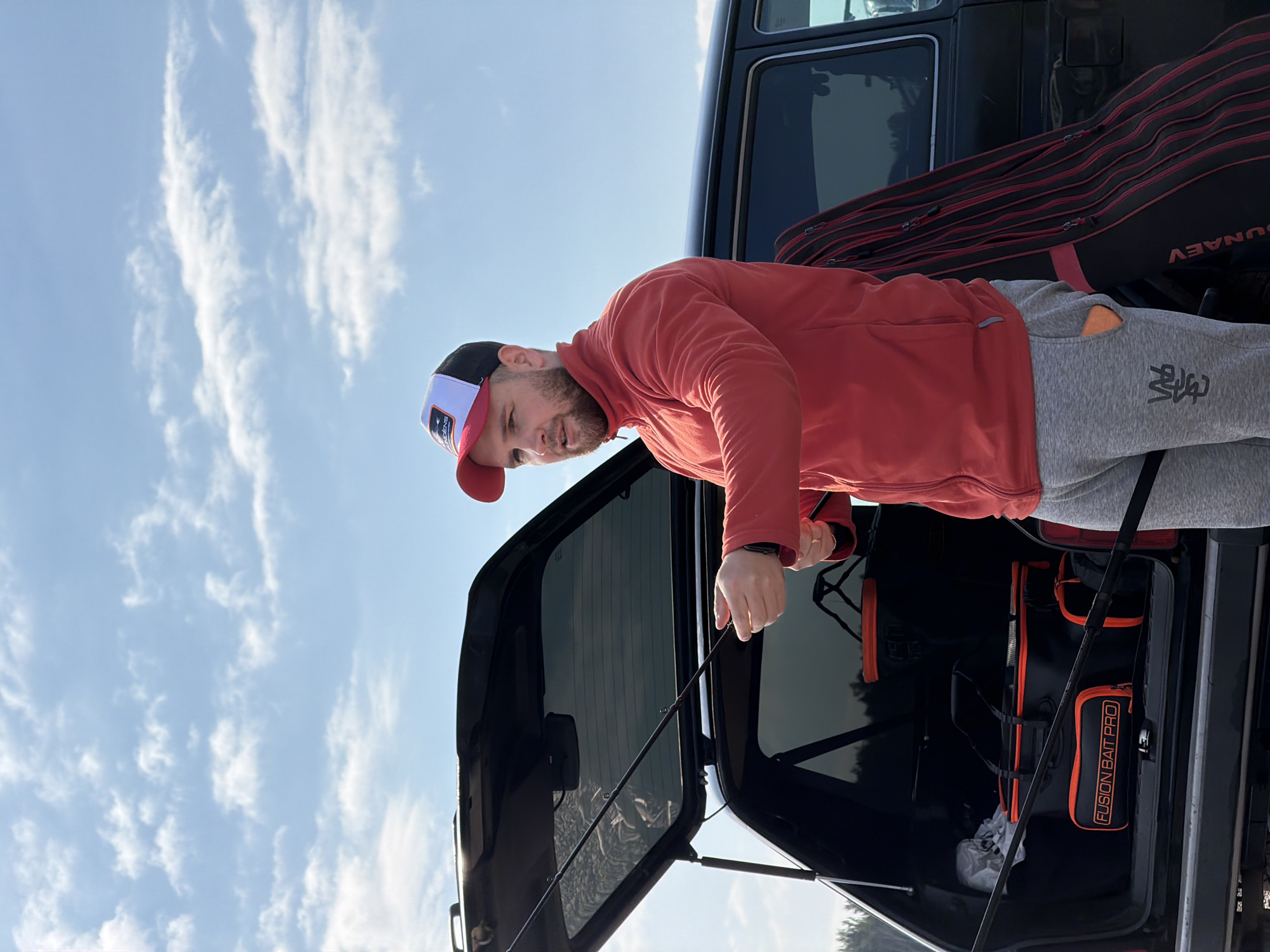 A man is standing at the open rear of a black SUV. He is wearing a red long-sleeve shirt, grey sweatpants with a logo on the left thigh, and a white, red, and black baseball cap. He is holding a fishing rod. The open rear of the SUV reveals black fishing equipment, including a bag labeled "FUSION BAIT PRO." A red and black fishing rod case is visible in the back of the SUV. The sky is visible in the background, with a light blue hue and scattered white clouds. The location is Chaykovsky, Russia.