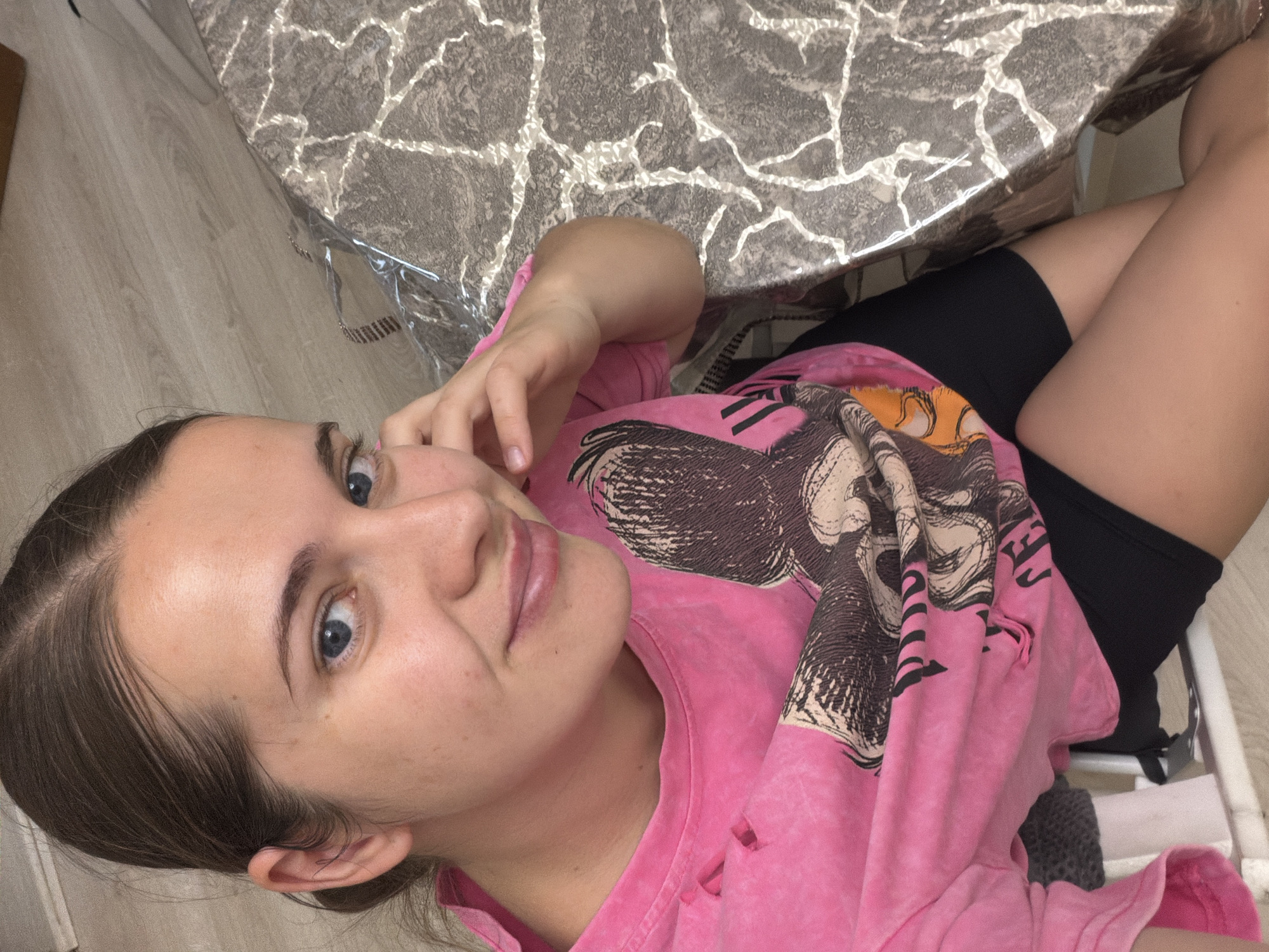 A young woman with blue eyes and light skin sits at a table. She is wearing a pink t-shirt with a large graphic design and black shorts. Her hair is pulled back. The table has a marble-patterned surface and is covered with a clear plastic protector. The floor is light-colored wood. The photograph was taken indoors. It is not possible to confirm the location is in Sochi, Russia, without additional evidence.