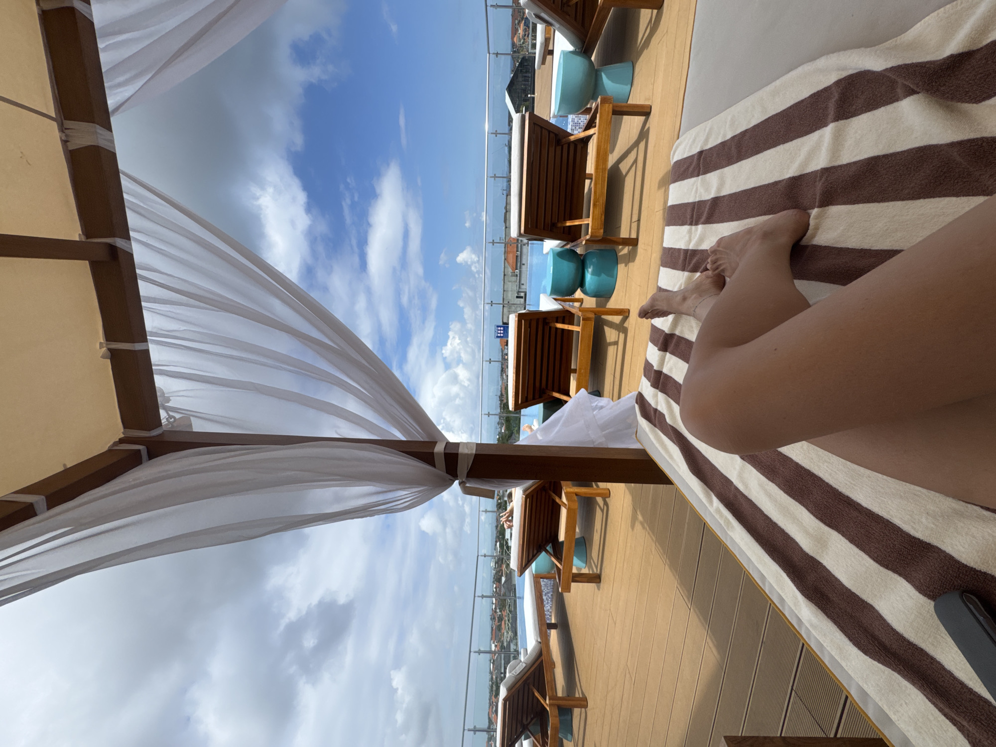 The image shows a person relaxing on a lounge chair under a canopy at a rooftop pool overlooking a city, likely in North Kuta, Indonesia. The person's legs and feet are visible, resting on a striped towel. Several other lounge chairs and bright blue cylindrical stools are arranged on a wooden deck beside a swimming pool. In the background, the city skyline with buildings and rooftops can be seen under a partly cloudy sky. White sheer curtains hang from the canopy, creating a partially enclosed, open-air seating area.