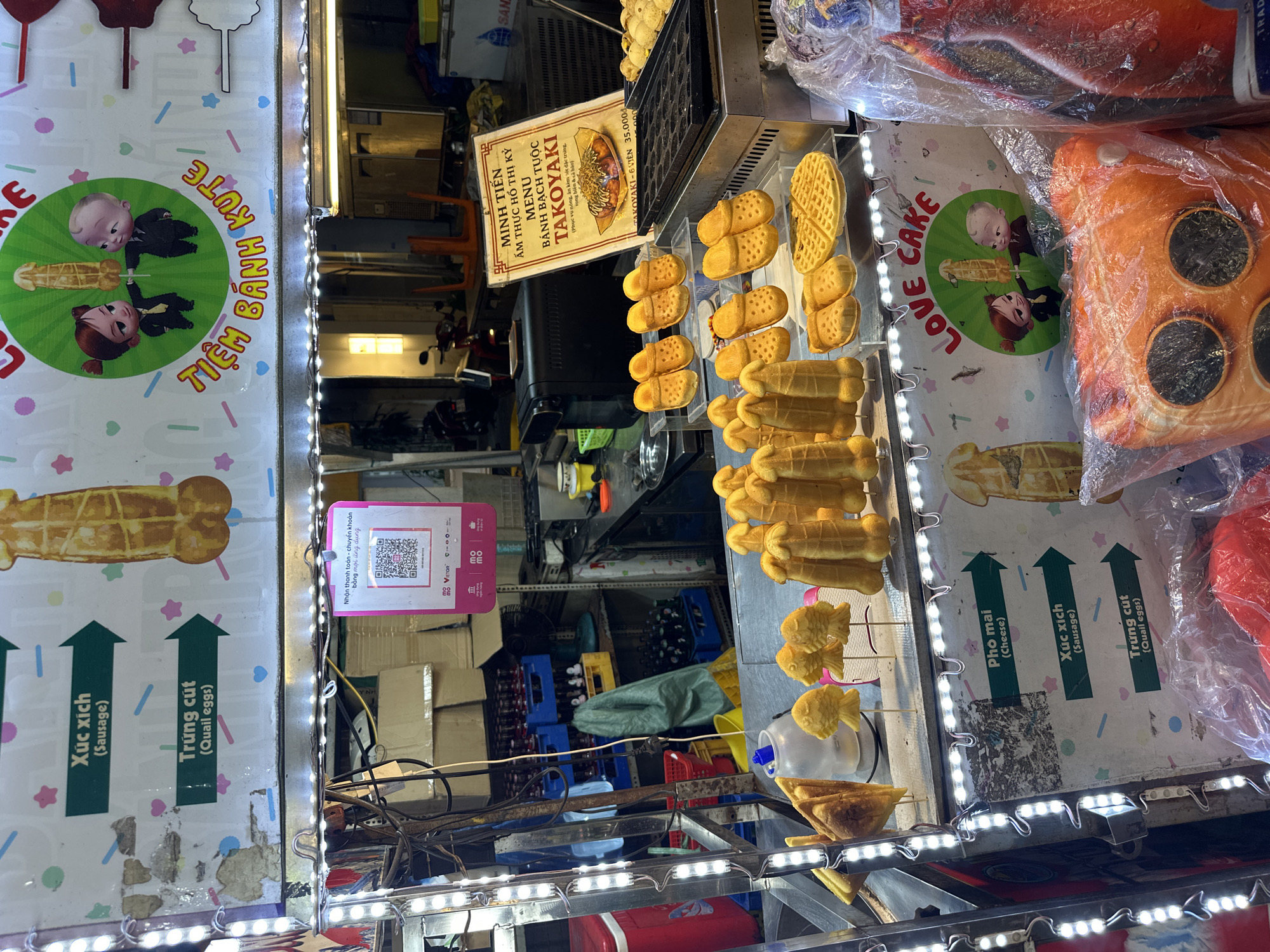 The image depicts a street food stall in Thủ Đức, Vietnam, featuring a variety of snacks. The stall is decorated with LED lights and signage written in Vietnamese. The primary food items displayed are waffles in different shapes (including fish and sausage shapes), takoyaki, and other baked goods. The stall's signage advertises takoyaki and provides prices. QR codes for payment are also visible. The setting includes a cluttered background of various boxes, bottles, and equipment associated with the food preparation and operation of the stall. A plush cushion resembling a pizza is seen on display. The signage includes cartoon figures of "The Boss Baby" and a little girl.
