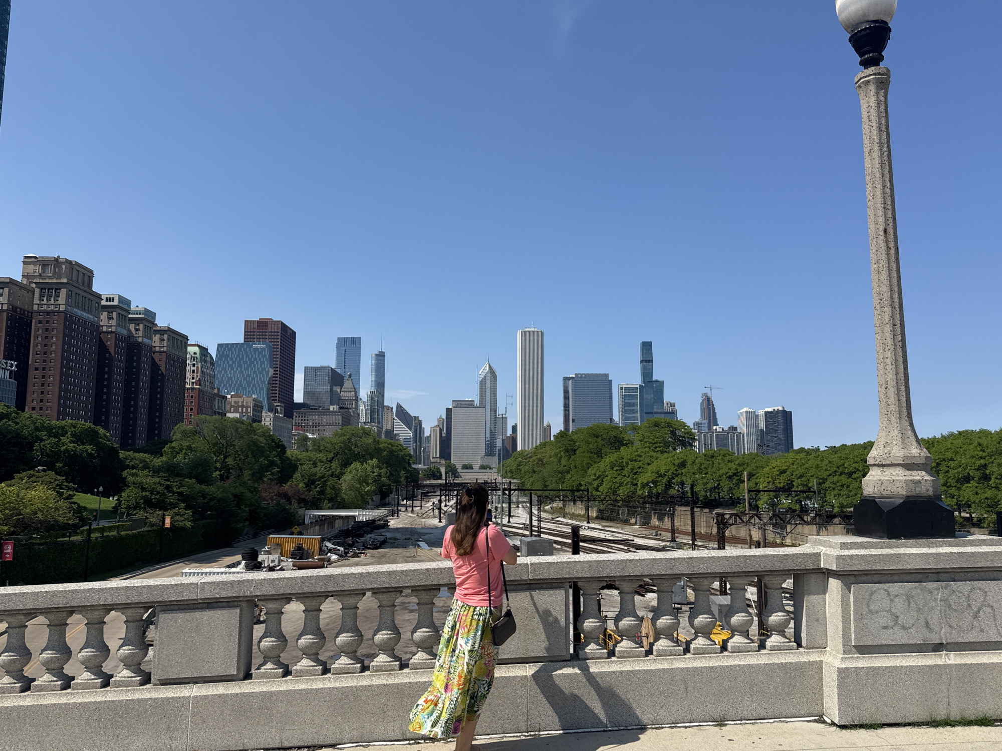 A full shot depicts a woman, viewed from behind, standing on a bridge. She is holding a smartphone and appears to be photographing the city skyline in the distance. She wears a pink shirt and a long, vibrant floral skirt. A black purse hangs from her left shoulder.
The bridge is constructed of grey stone and features a decorative railing. A tall, ornate lamppost stands to the right of the frame.
In the background, a sprawling cityscape is visible. Numerous skyscrapers of varying heights and architectural styles dominate the horizon. The area between the bridge and the city buildings is partially obscured by greenery and what appears to be railway tracks.
The scene is set on a bright, sunny day, with a clear, blue sky overhead. The image is taken in Chicago, Illinois, United States of America, likely from a bridge in Grant Park, looking towards the central business district, commonly referred to as "The Loop".