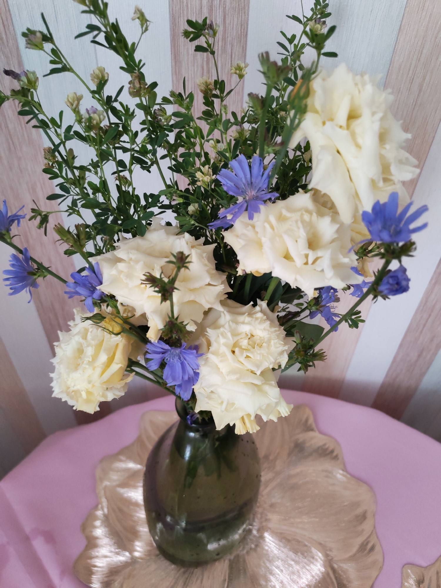 The image displays a bouquet of cream-colored roses and small blue flowers, possibly chicory, arranged in a dark green glass vase. The vase sits on a textured, light-colored placemat, which is on top of a pink tablecloth. The background consists of a wall with vertical stripes of light beige and white. The scene is indoors, with no visible indications of a specific location within Izhevsk, Russia. There are no people or animals present in the image, and no actions or interactions are depicted. The lighting appears to be natural, casting soft shadows. The composition focuses on the floral arrangement and the immediate surface it rests upon.