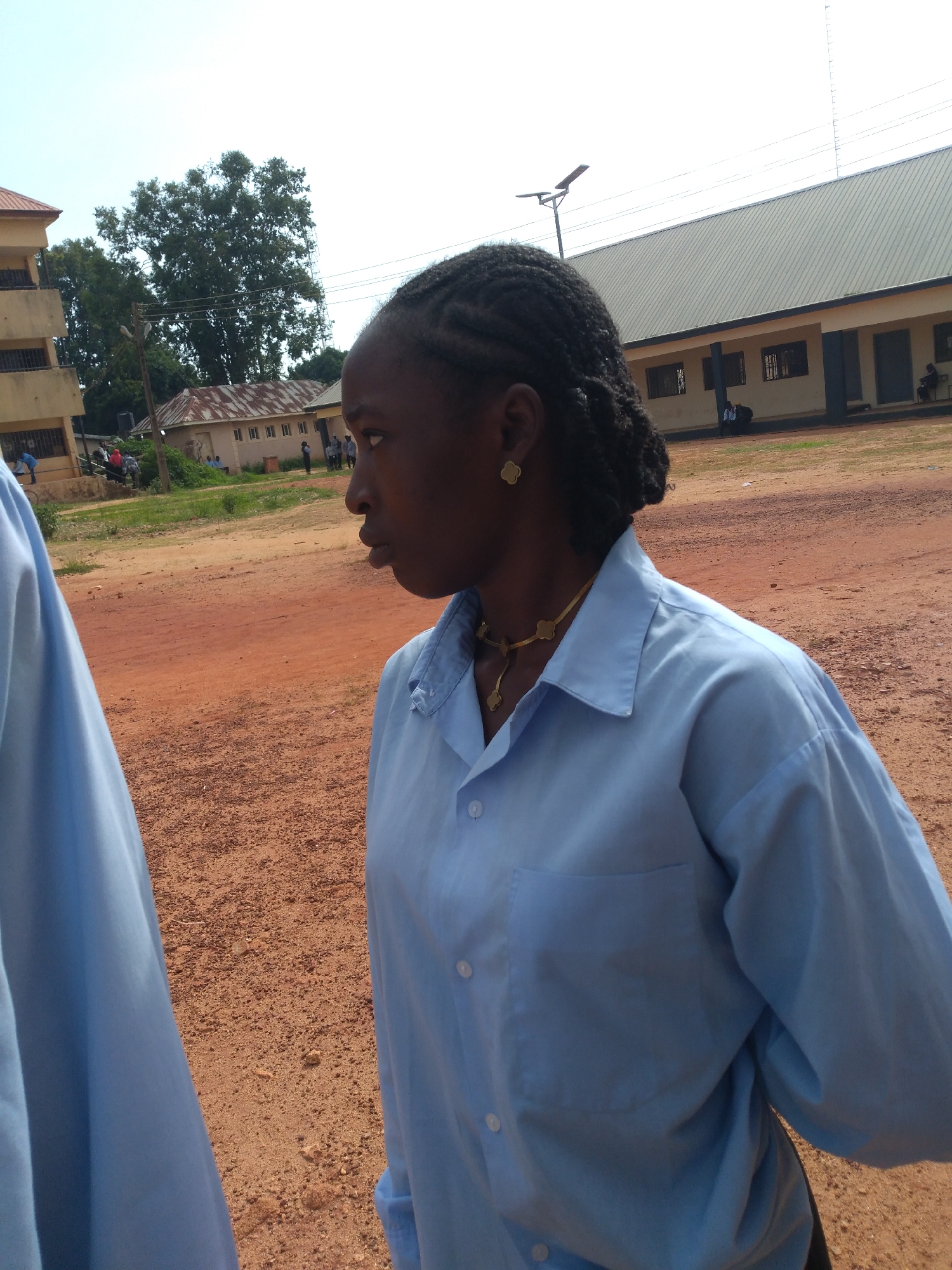 The image depicts an outdoor scene, possibly on the grounds of a school or administrative building in Jos, Nigeria. In the foreground, a young woman of African descent is shown from the chest up, facing to her left in profile. She has her hair in braids and is wearing a light blue collared shirt and gold jewelry, including earrings and a necklace. The texture of her hair and the detail of her jewelry are visible. To the left of the frame, a portion of another person wearing a similar light blue shirt is visible, also in profile.
The background features a dirt or gravel ground, characteristic of many unpaved areas in Jos. Behind the subjects, there are several low-lying buildings with beige walls and corrugated metal roofs, suggesting a utilitarian architectural style common in the region. One building appears to be a two-story structure with visible windows. In the distance, a large tree stands prominently against a bright sky. Power lines and a solar-powered street light are also visible, indicating the presence of infrastructure. A few other individuals and some parked vehicles are discernible in the background, hinting at a public or communal space. The overall lighting suggests it is daytime, possibly late morning or early afternoon, with no strong shadows present.