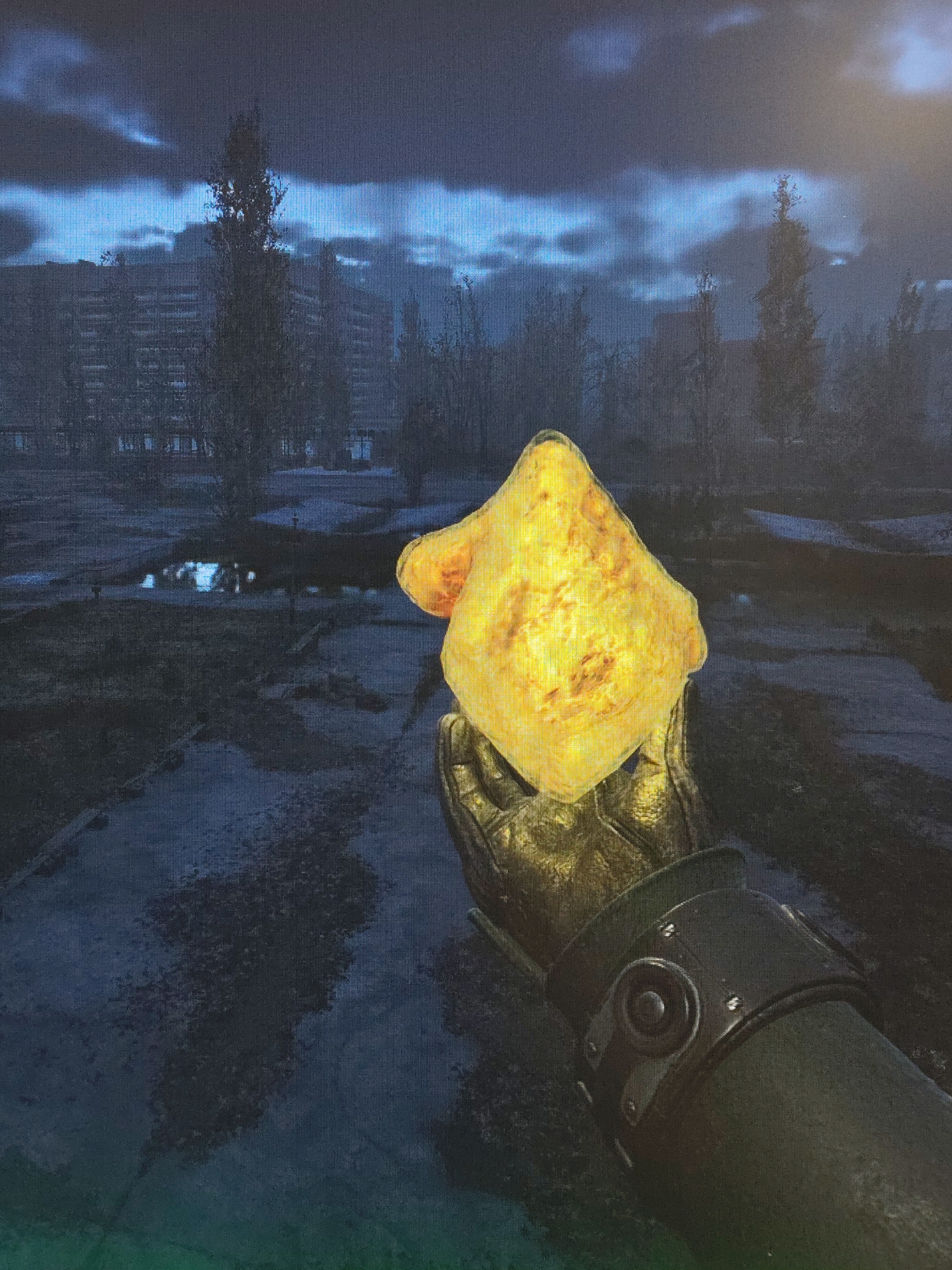 The image displays a first-person perspective from a video game, showing a gloved hand holding a brightly glowing, irregularly shaped, yellowish object against a bleak, desolate background. The hand is covered by a dark, textured glove, possibly leather or heavy fabric, with a metallic-looking, armored cuff on the wrist.
The object held is the focal point, emitting a strong yellow-orange light that illuminates parts of the glove. Its shape is amorphous, resembling a mineral formation, a strange artifact, or perhaps an unnatural, luminous food item.
The background depicts an abandoned urban or semi-urban environment under a dark, overcast sky with hints of blue light peeking through the clouds. Silhouettes of bare, spindly trees are scattered across the landscape, and in the distance, dark, multi-story buildings with visible windows are visible, suggesting derelict structures. The ground appears uneven and dark, with reflections from what could be puddles, mirroring the dim sky.
The overall atmosphere is dark, somber, and mysterious, typical of a post-apocalyptic or survival-horror game setting. The image itself appears to be a photograph of a digital screen, as indicated by a noticeable grid-like pixelation pattern across the entire frame. No text is visible in the image.