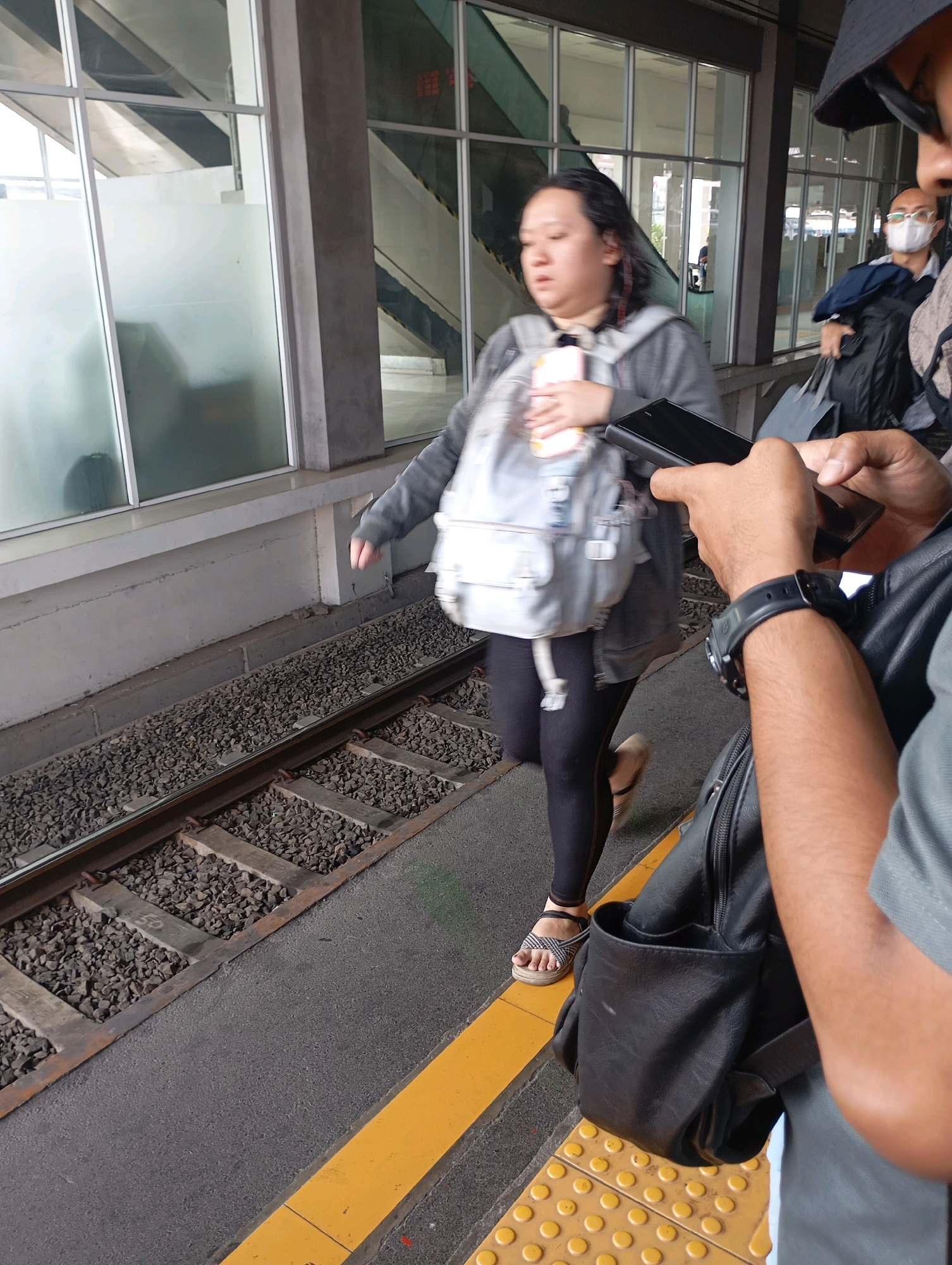 An Indonesian woman with a backpack runs alongside train tracks at a train station in West Jakarta, Indonesia. She is wearing black leggings, a grey hooded jacket, and sandals. The woman is holding a pink object in her right hand and a white and pink item in her left hand. A man in the foreground is holding a black smartphone. In the background, another man wearing a white face mask is walking away from the camera, carrying a dark bag. The train tracks are made of gravel and wooden ties. The platform has a yellow tactile warning strip. The scene appears to be in an outdoor, covered area of the train station, with large windows and concrete pillars visible.