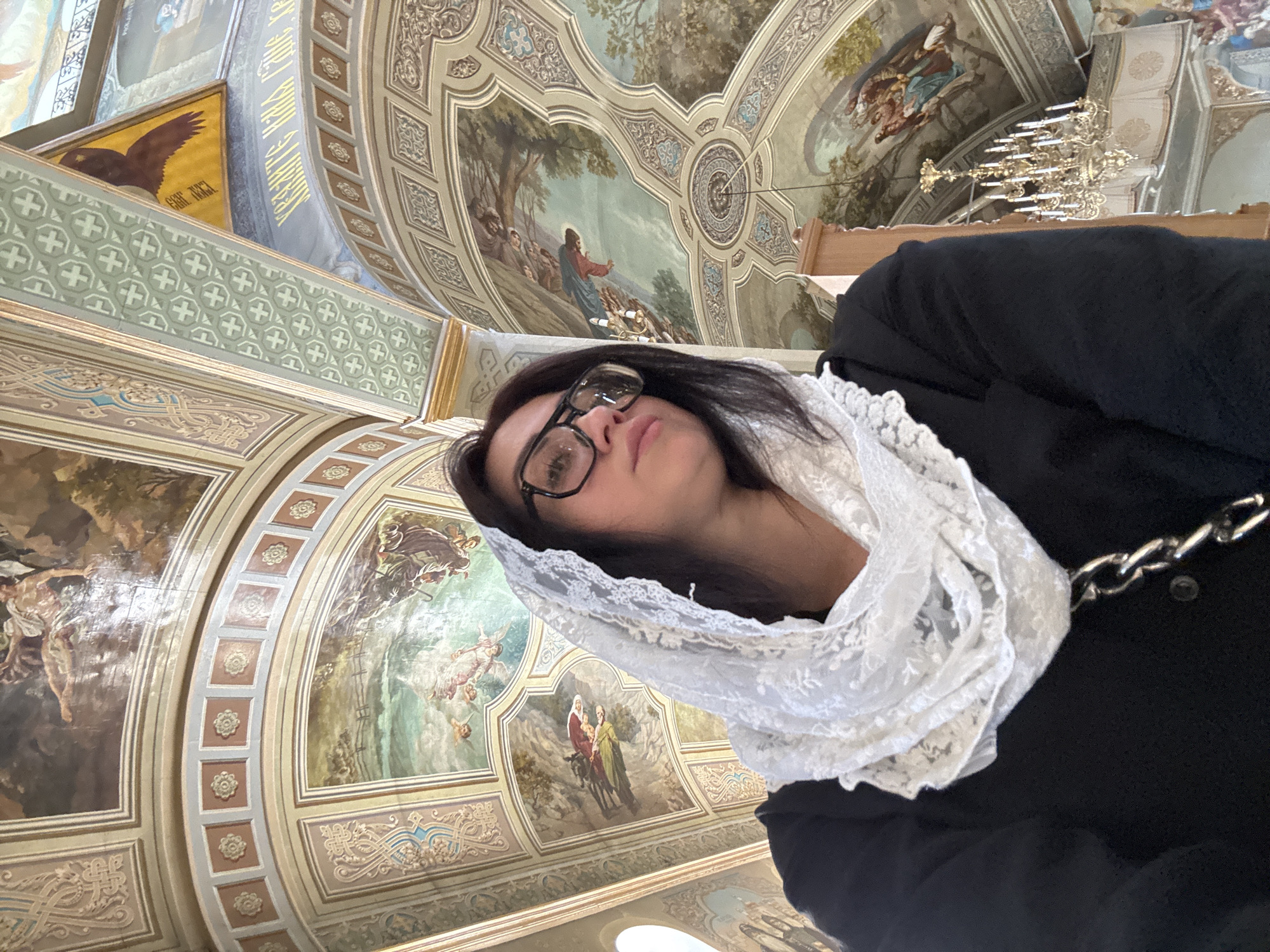 An individual is taking a selfie indoors, positioned in the foreground. They are wearing a black garment and a white lace headscarf, with large-rimmed glasses on. The background is an elaborately decorated ceiling with frescoes and architectural elements. The ceiling features religious imagery, including depictions of figures and ornate patterns. A chandelier is visible in the upper right portion of the image, contributing to the interior ambiance. The location is not specified as being in a particular city.