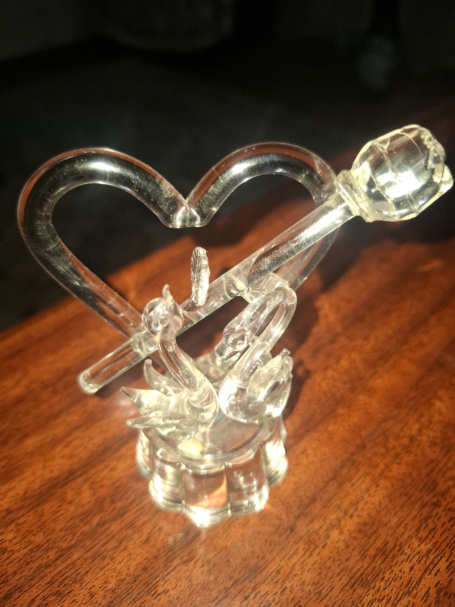 A clear glass sculpture of two swans forming a heart shape is displayed on a wooden table. The sculpture is illuminated from the side, casting shadows. The background is dark and out of focus. The table has a polished wood grain. The sculpture features a heart shape formed by the swans' necks and bodies, with a faceted glass element at the top right resembling a flower or crystal. Two swan heads are visible, facing each other. The base of the sculpture is circular and ribbed. The image appears to be taken indoors, possibly in a home or display setting. There are no people present in the image, and no specific location in Almaty, Kazakhstan can be identified from the visual information provided.