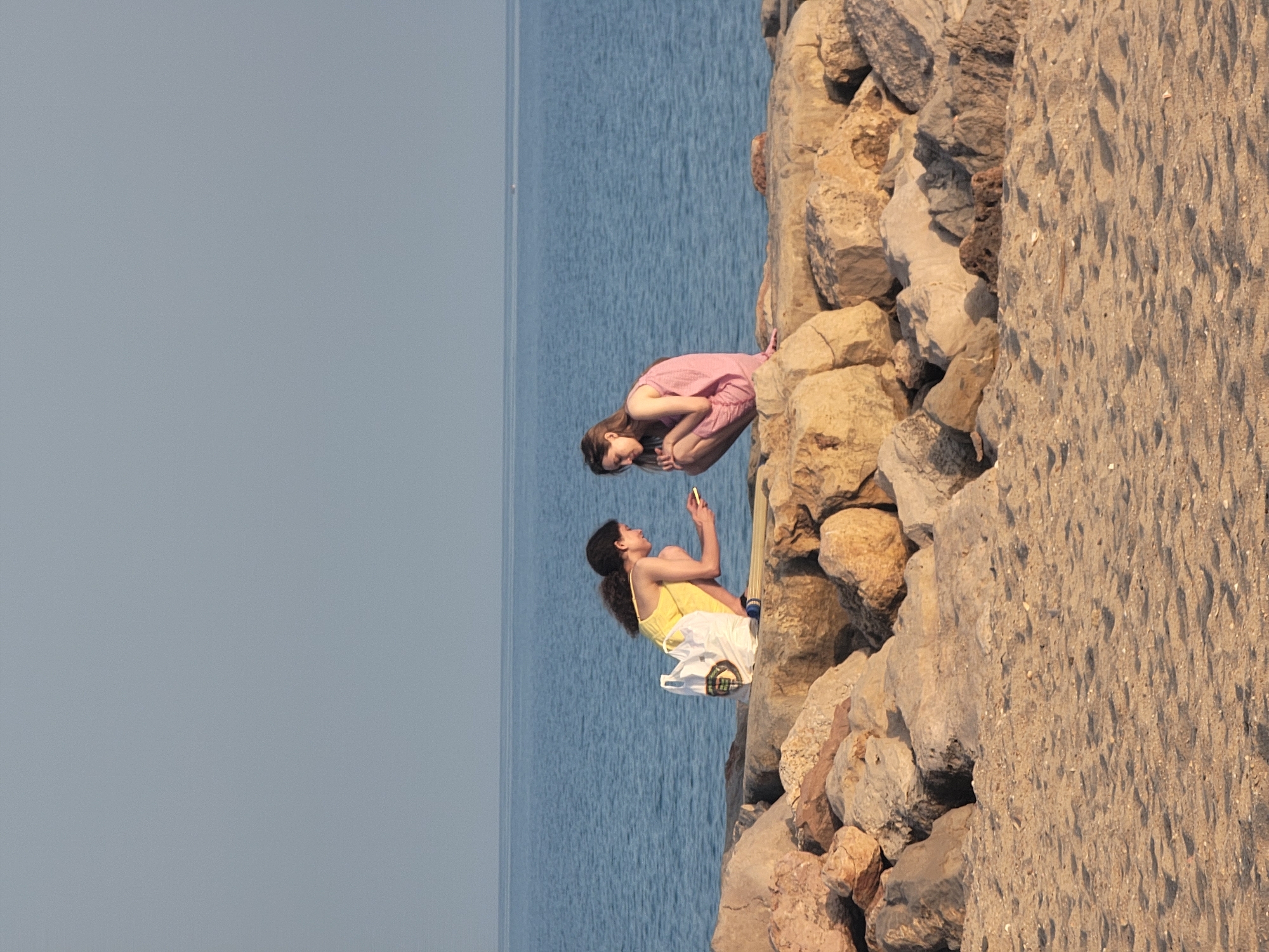 Two young women are crouched on a rocky breakwater near the beach in Quarteira, Portugal. The woman on the left, wearing a yellow sundress, is looking at her phone, which she holds in her hand. A white canvas bag with a checkered pattern rests beside her. The woman on the right, in a pink gingham dress, is looking down, her knees drawn up to her chest. The ocean is calm and blue in the background, with a clear sky above. The breakwater is made of large, rough-hewn rocks, and the sandy beach is visible in the foreground. The scene suggests a relaxed, warm day by the sea.