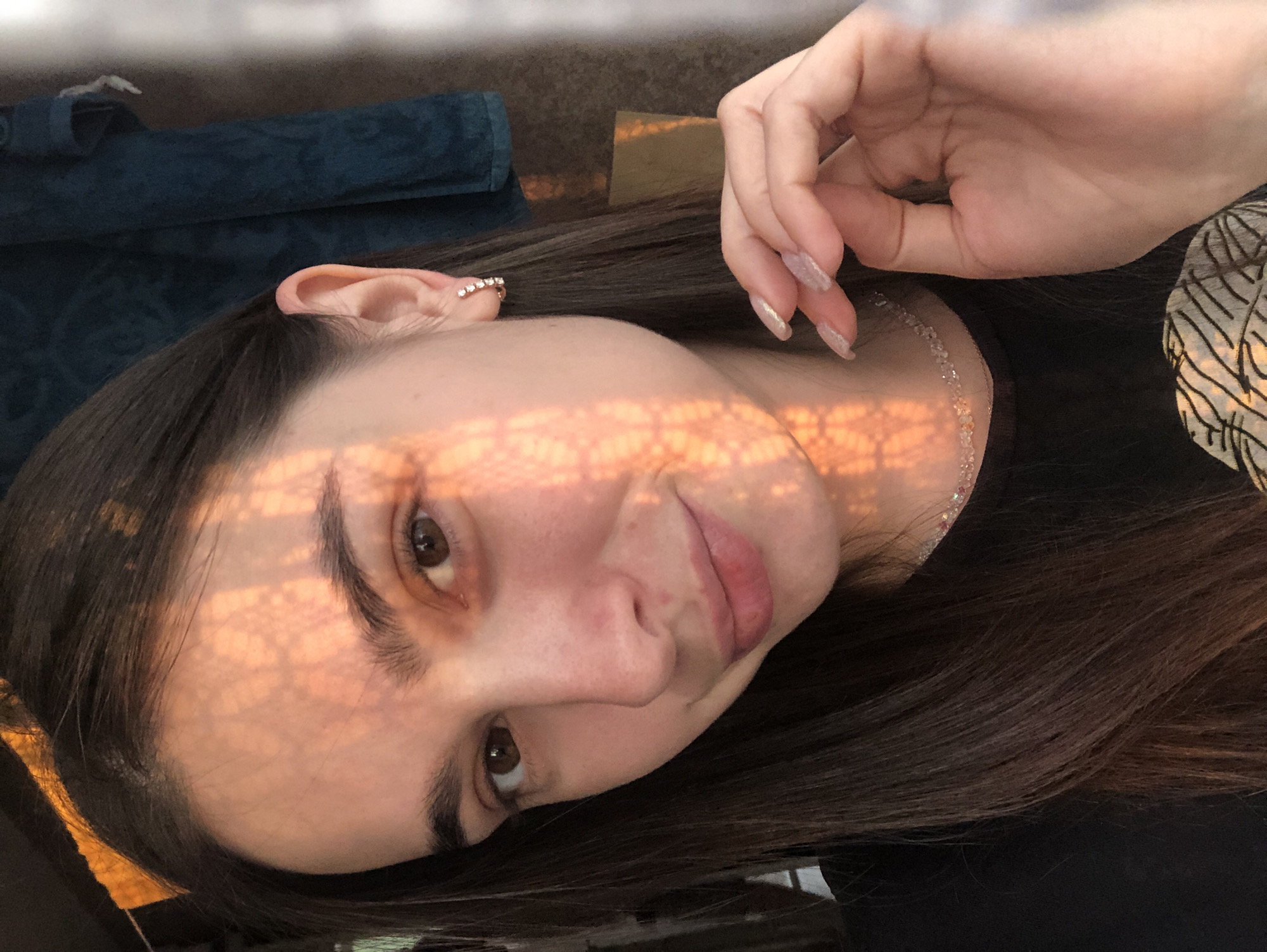 A young woman with long, dark brown hair is captured in a selfie. She is wearing a black t-shirt and a delicate necklace. Her right ear, adorned with a small hoop earring featuring several clear stones, is visible. The woman's hand, with its nails painted in a pearlescent shade, is positioned near her face and neck. A patterned lace shadow is cast across the left side of her face and forehead, suggesting the presence of sunlight filtering through a patterned object. The background is somewhat out of focus, showing a dark teal fabric, possibly a towel or curtain, hanging to the right, and a wooden surface on the far left. The lighting indicates it is likely daytime, with warm, golden sunlight illuminating the scene. The woman is looking directly at the camera with a slight smile. The image was taken in Chaikovsky, Russia.