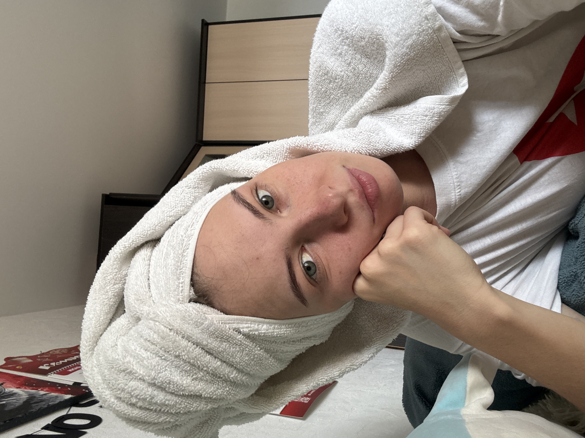A young woman with a white towel wrapped around her head and shoulders poses for a selfie indoors. She is wearing a white t-shirt with a red star design on it. Her right hand is resting on her chin. In the background, a white wall with posters and a closet with wooden doors are visible. The lighting suggests it is daytime, possibly in a bedroom or living area. The scene is static and appears to be a casual, personal moment. The location is identified as Perm, Russia.