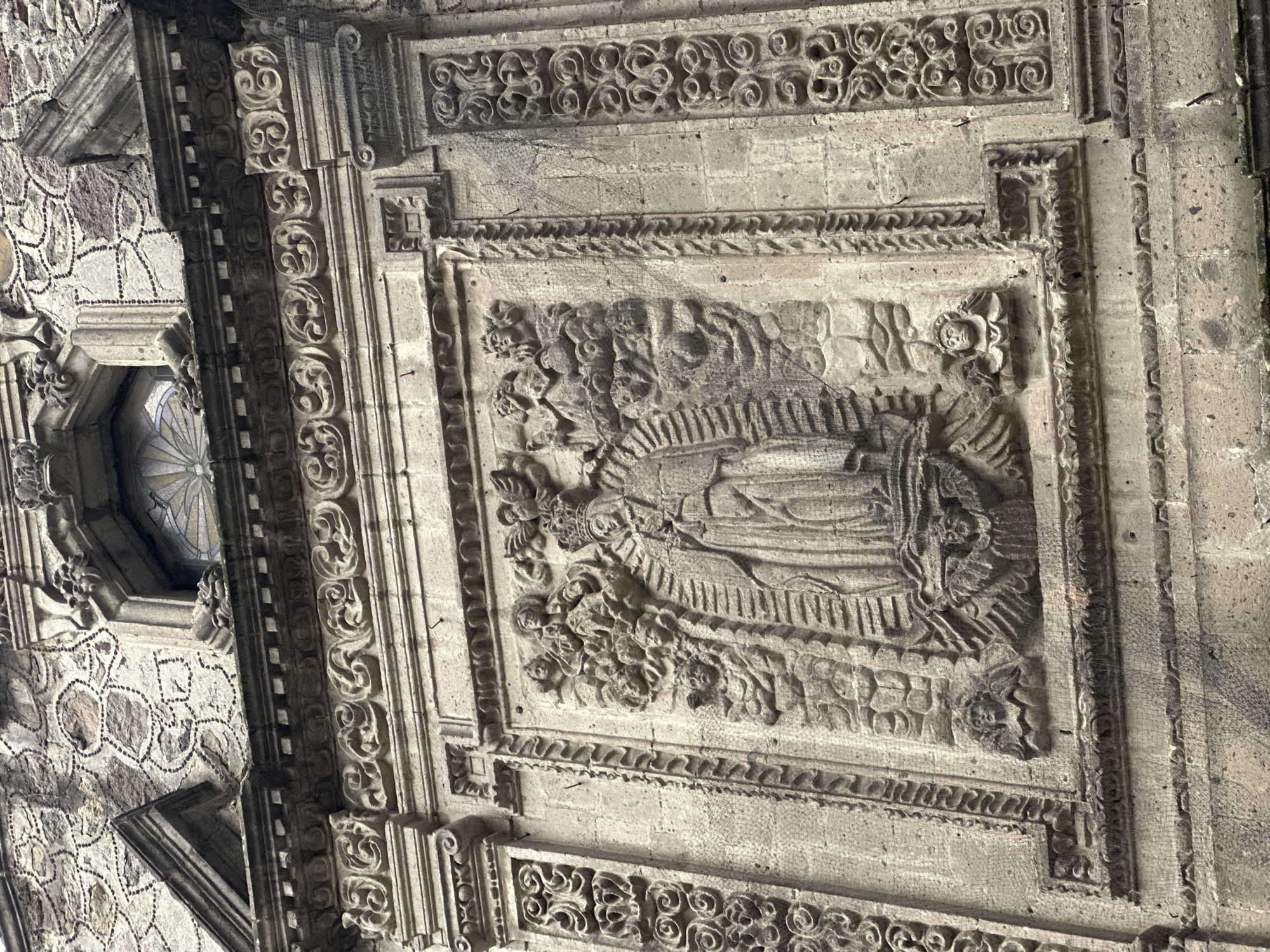 The image shows a close-up of architectural details on a stone facade, likely belonging to a building in Mexico City, Mexico. A rectangular stone carving with a religious scene dominates the center. The carving depicts a figure resembling the Virgin Mary, encircled by floral motifs and what appear to be winged angels. The carving is framed by layers of decorative stone elements, including geometric patterns, scrollwork, and carved borders. To the left of the carving, there is a section of a wall constructed from irregularly shaped stones with mortar. A window with a multi-pane design is positioned at the upper left. The overall tone of the image is aged and textured due to the stone material and possible weathering.