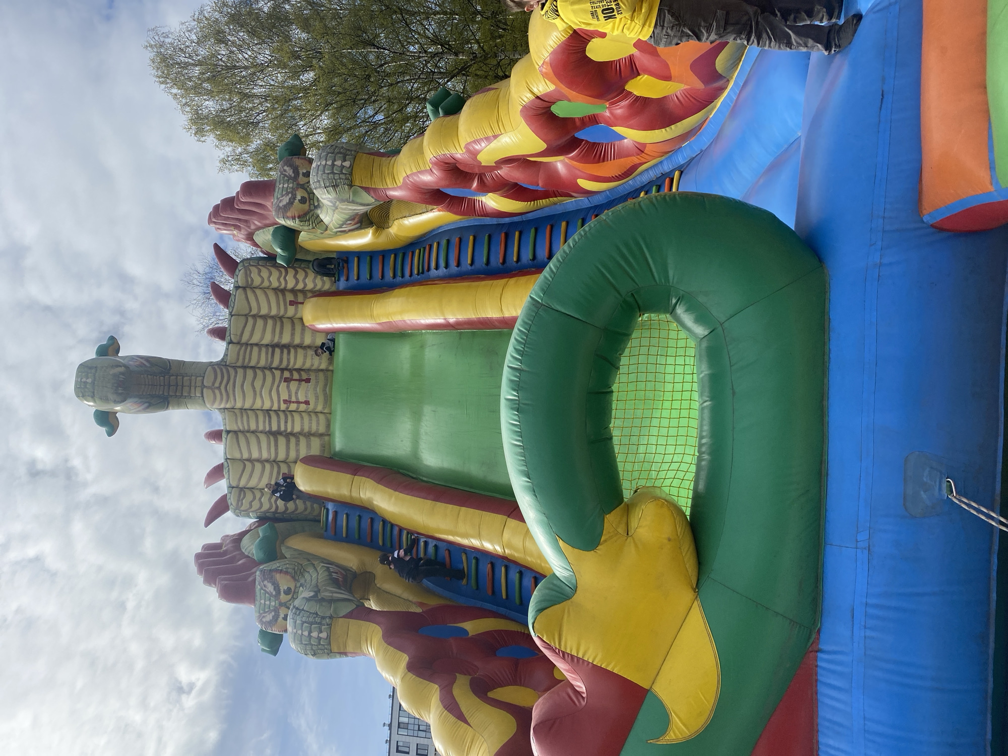 The image shows a large inflatable dragon-themed bouncy castle, likely set up for a children's event. The structure is multicolored with green, yellow, red, blue, and tan sections and depicts dragon heads and scales. A green inflatable ring is attached to the side. A person in a yellow shirt is partially visible at the top of the frame.
The surrounding environment includes a cloudy sky and trees in the background, along with a building. The exact location in Saint Petersburg, Russia cannot be confirmed with the available details.