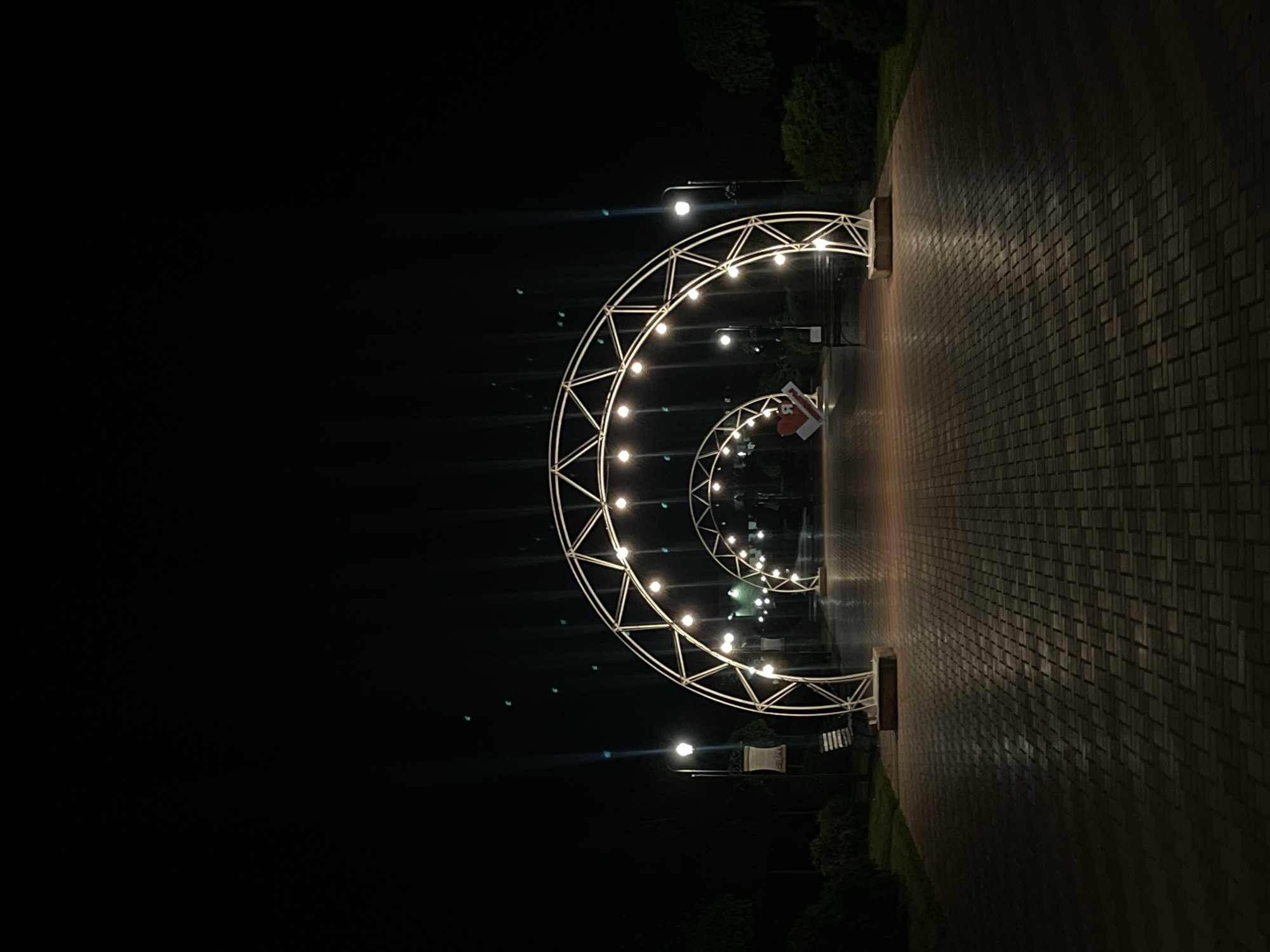 The image shows a brick walkway at night in Bender, Moldova. The walkway is lined with a series of lighted, semi-circular metal arches, creating a tunnel effect. Streetlights are visible, and there is vegetation bordering the walkway. A sign with a red heart is visible within the arches. The right side of the image shows the walkway texture. The left side of the image is dark.
