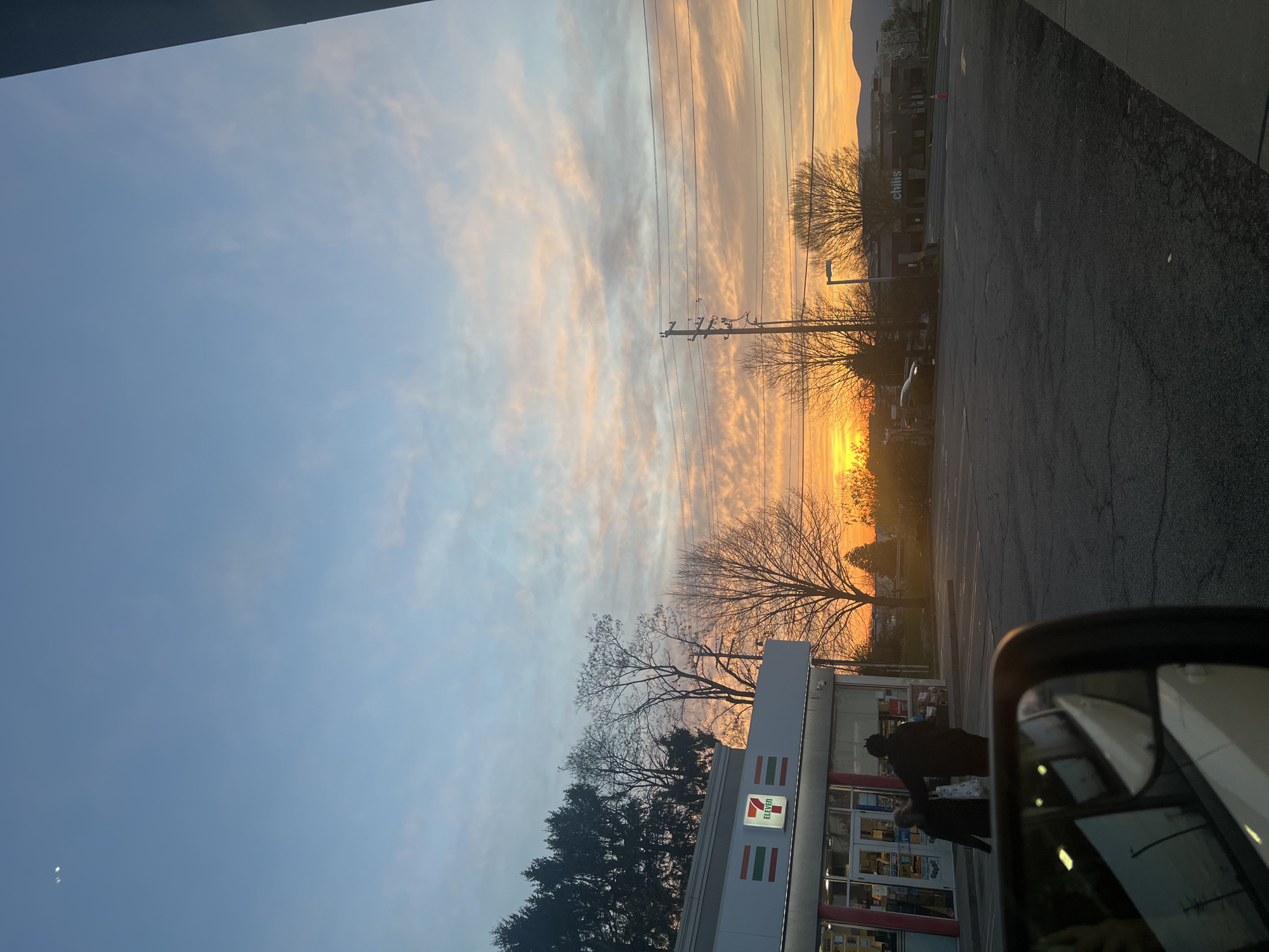 The image captures a sunset sky over a 7-Eleven convenience store in the City of Newburgh, United States of America. The sky is colored with hues of orange and blue. Power lines run across the view. A parking lot is visible, and individuals are present near the store entrance. A side mirror of a car is visible in the foreground. In the background, a building labeled "Chili's" can be seen.