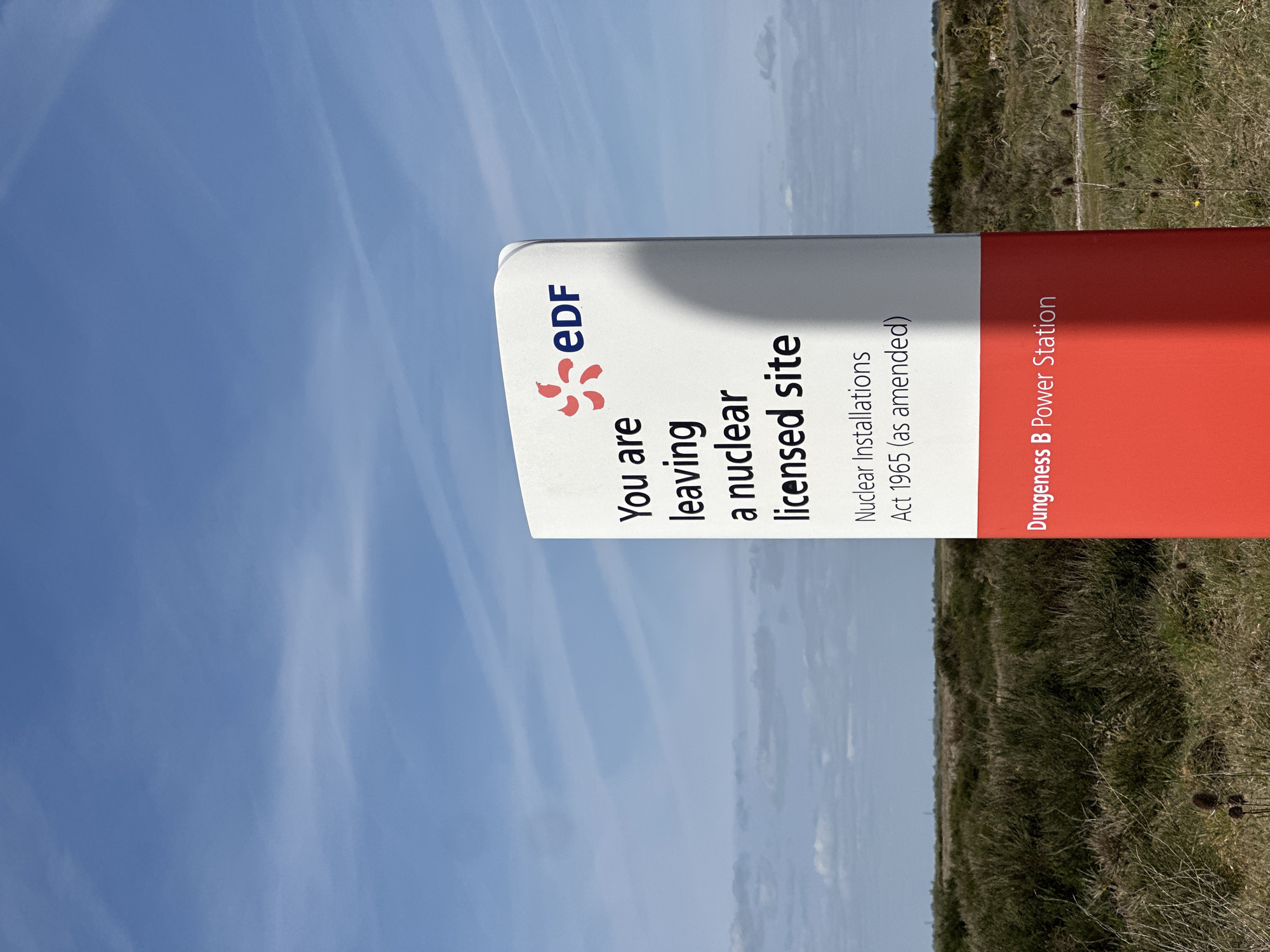 The image depicts a sign indicating the boundary of a nuclear licensed site, belonging to EDF. The sign reads "You are leaving a nuclear licensed site. Nuclear Installations Act 1965 (as amended). Dungeness B Power Station." The sign is white on the upper portion with the EDF logo, transitioning to red at the bottom with the power station name. The background features a blue sky with wispy clouds, and a field with vegetation, likely located near Dungeness B Power Station. Given the location information, this is most likely in the Folkestone and Hythe District, United Kingdom.