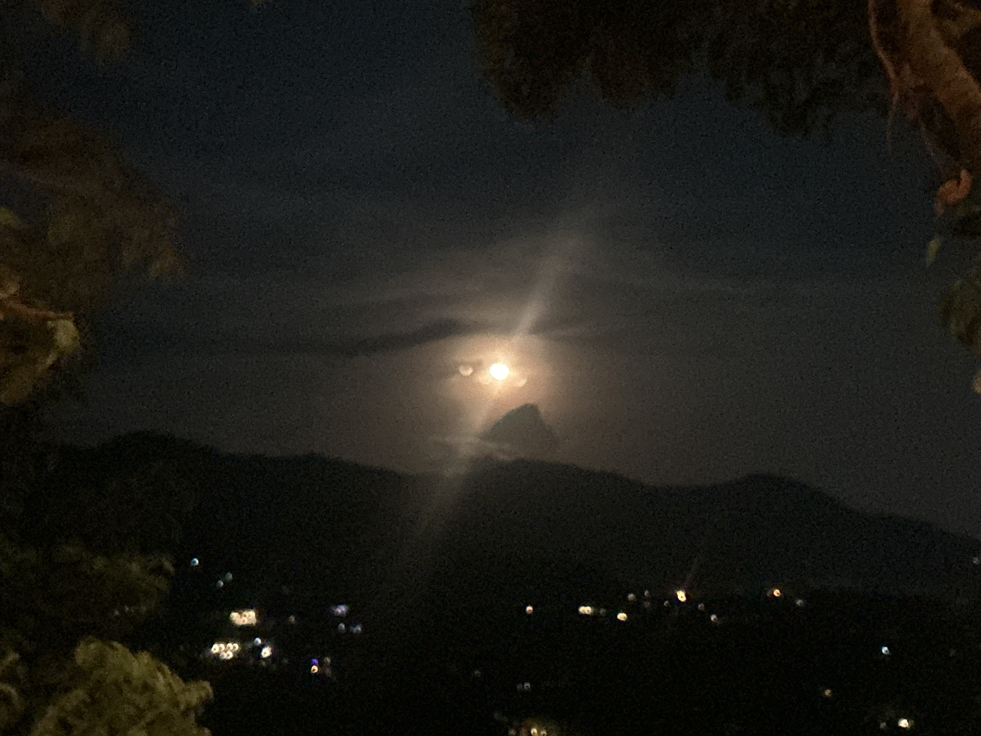 The image depicts a night scene in Baan Hin Kong, Thailand. A bright moon is visible in the sky, partially obscured by clouds. Below the sky, a silhouette of mountains is seen, with scattered lights suggesting a populated area. The foreground includes foliage, partially framing the view. The image appears to be taken from an elevated position.